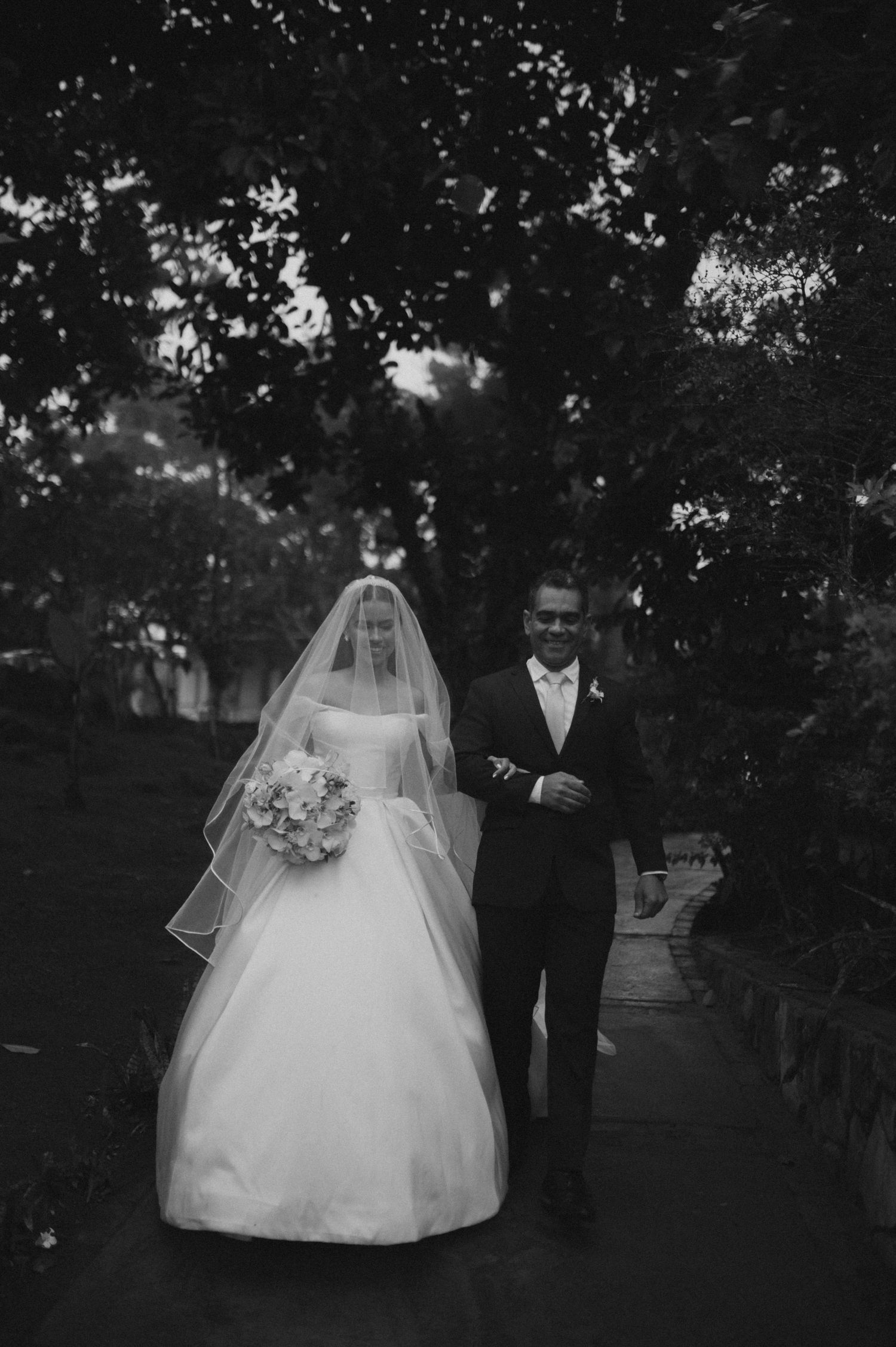 A black and white wedding photo showing a couple walking along a tree-lined path in formal attire.