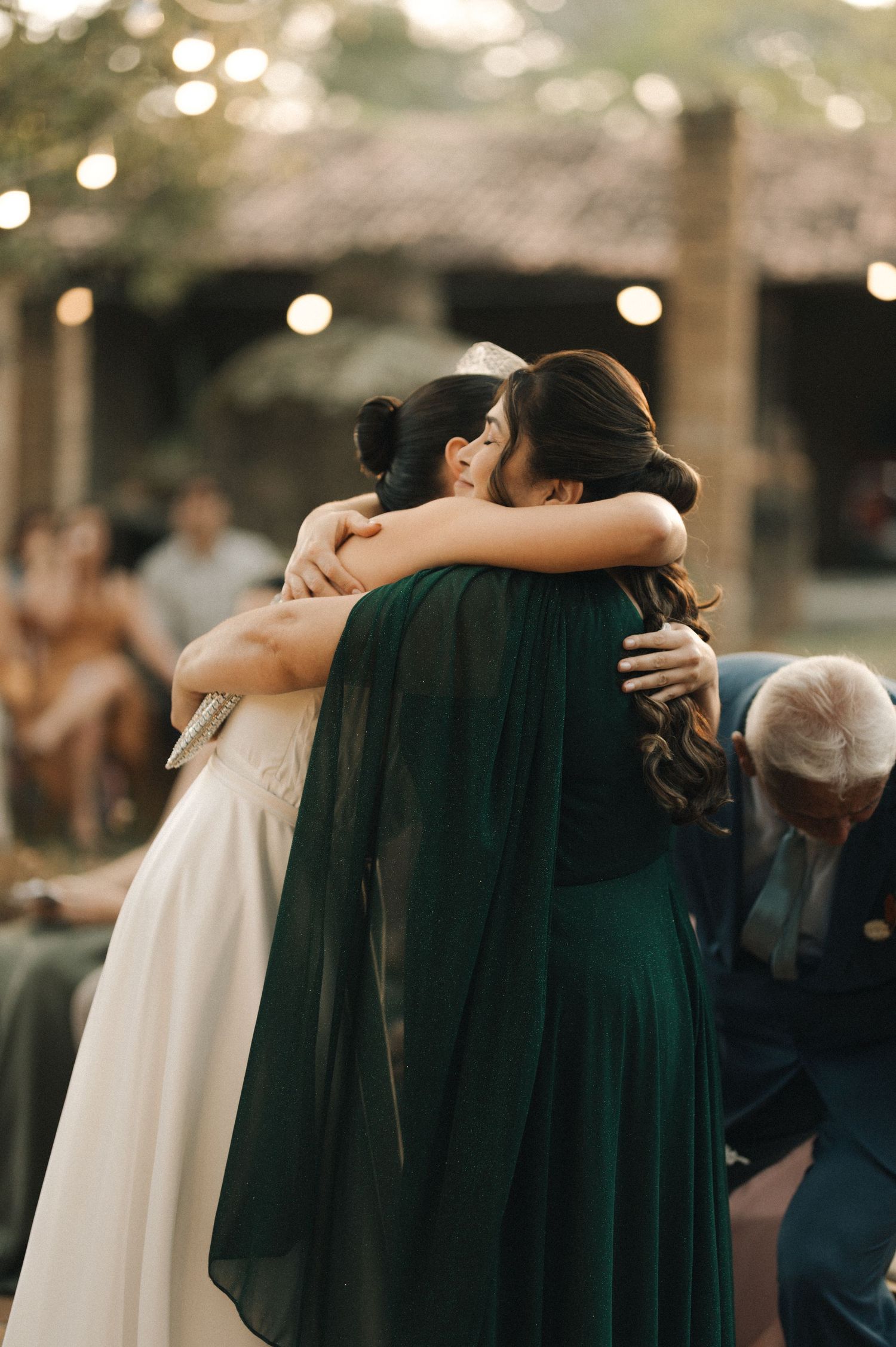 Two people share an emotional embrace at a wedding, with one wearing a white gown and the other in emerald green velvet.