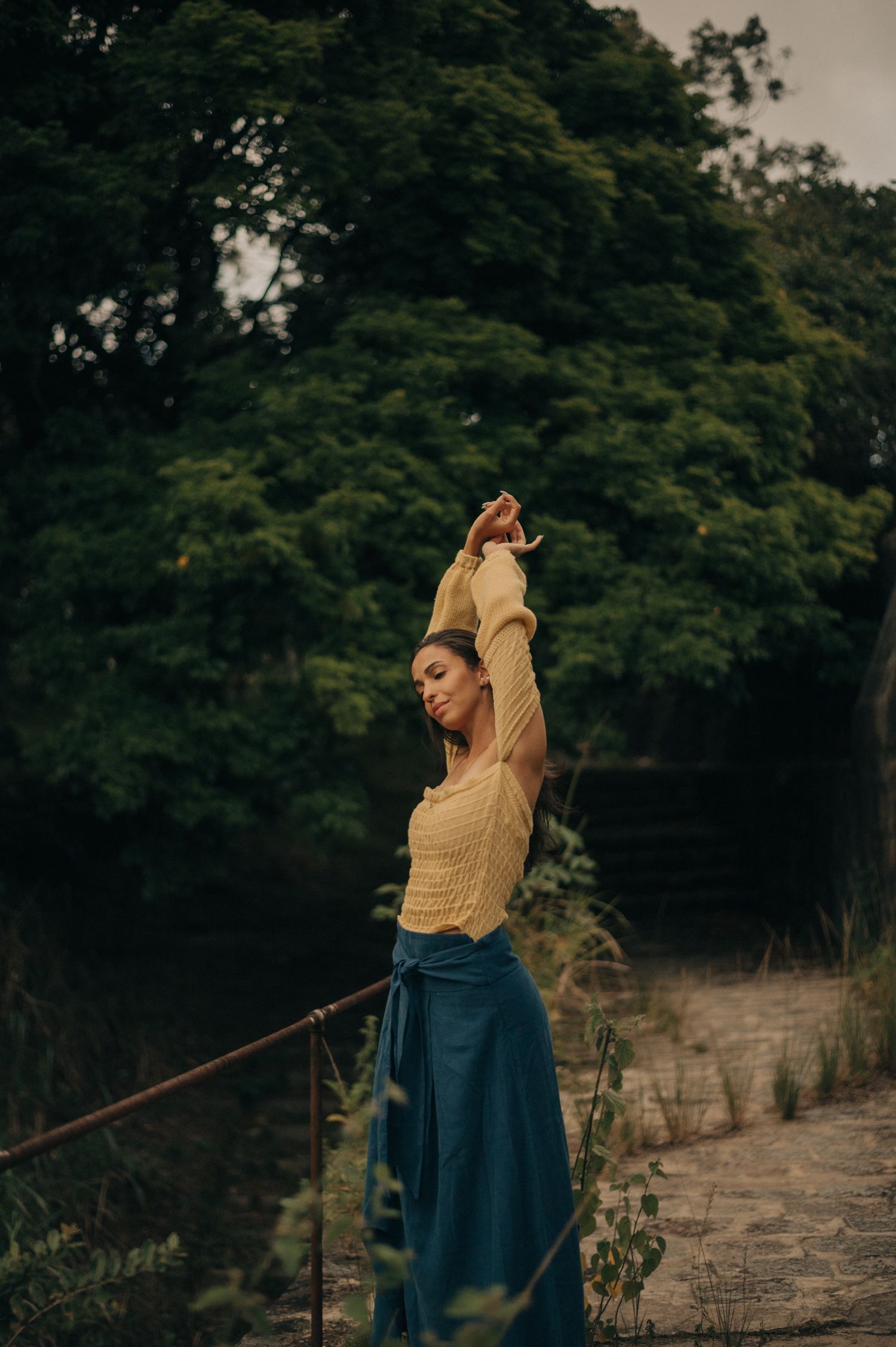 A figure in yellow top and blue skirt stretches gracefully against a backdrop of lush trees.