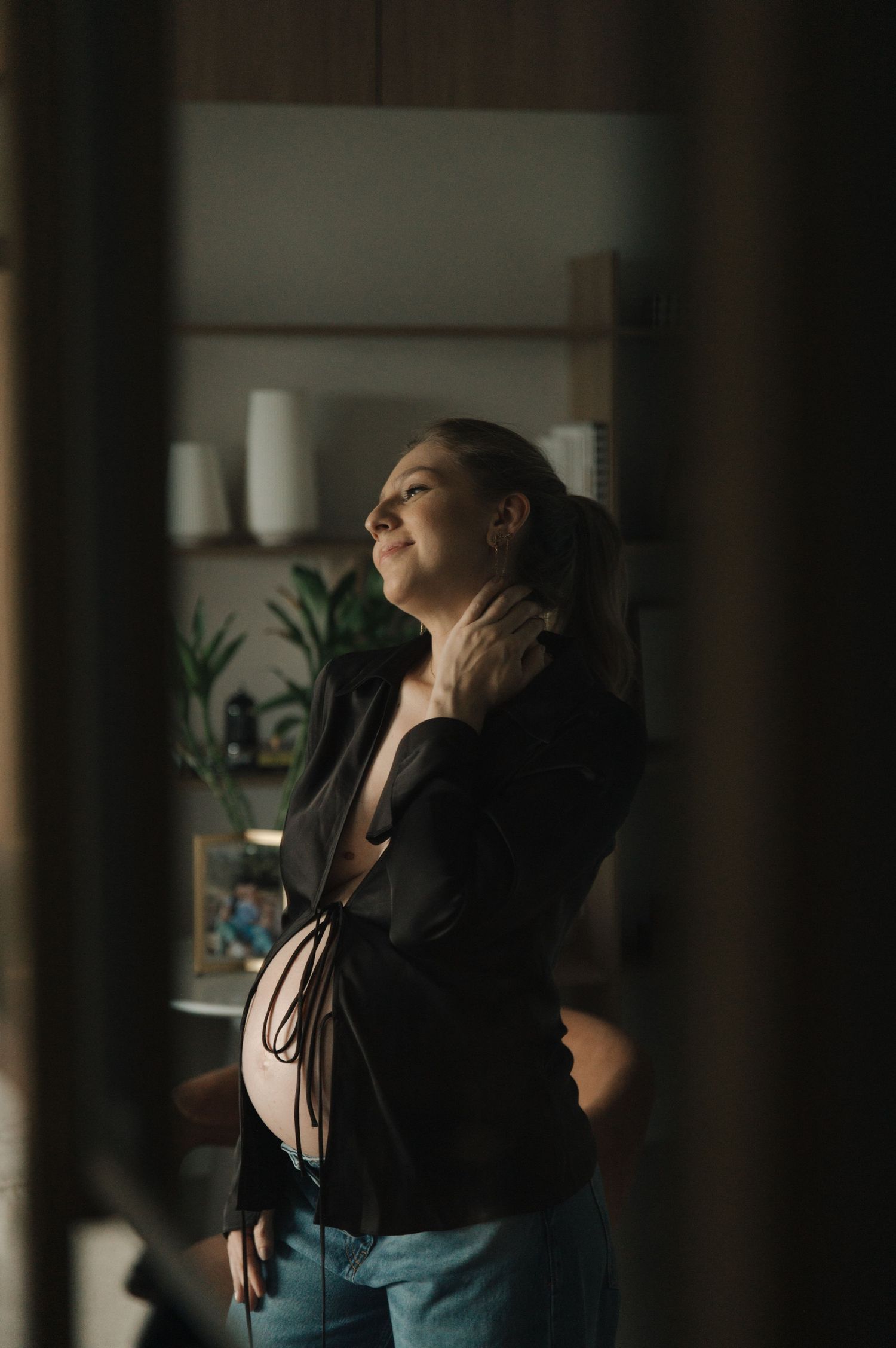 Candid maternity moment captured in moody lighting with indoor plants in background.