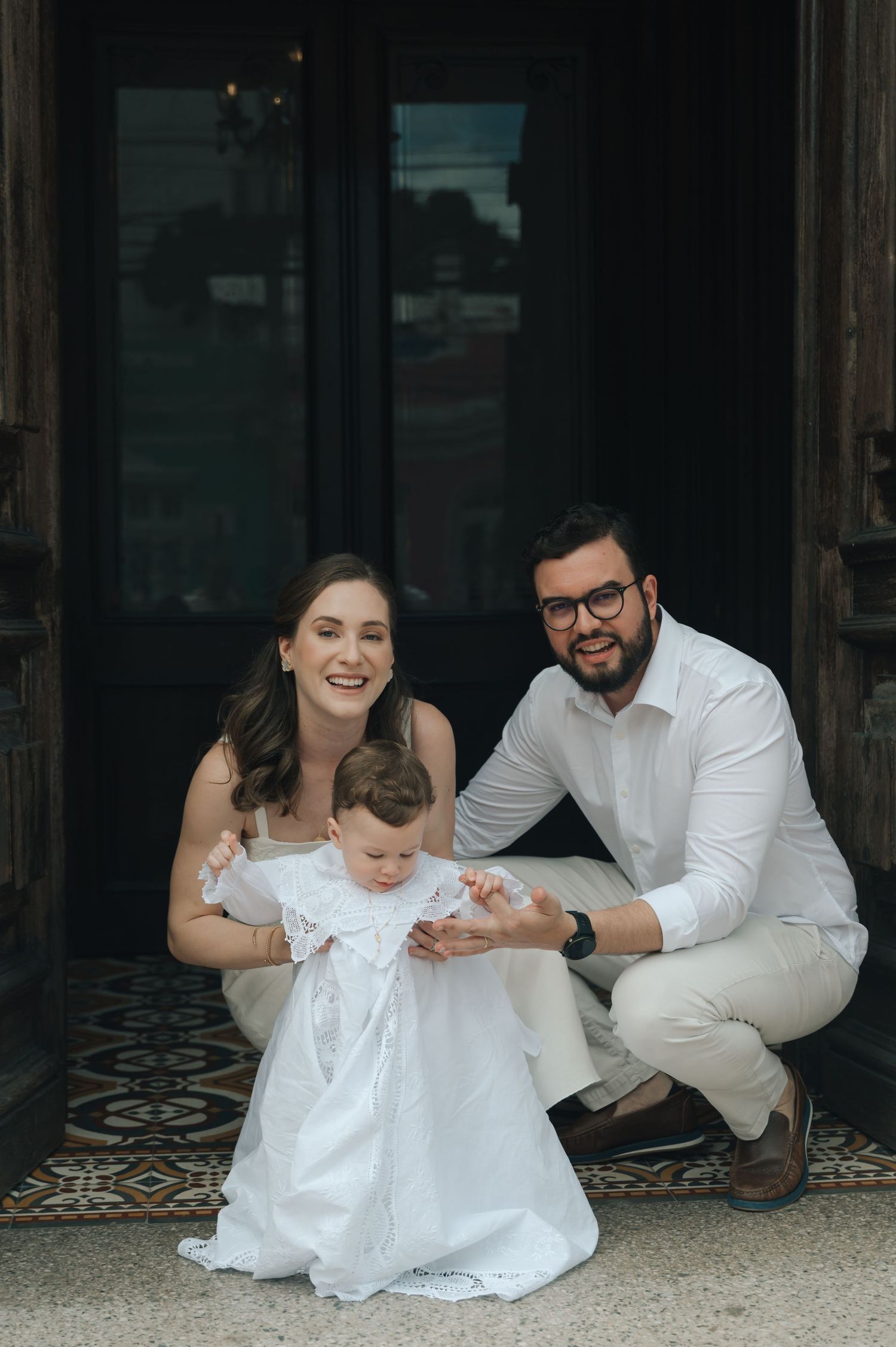 A family in white clothing sits together on a patterned floor during a photo session.