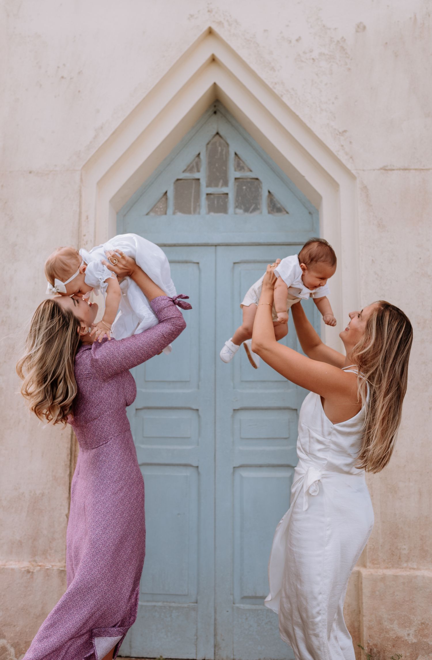 Two people in purple and white dresses dance playfully in front of a light blue vintage door.