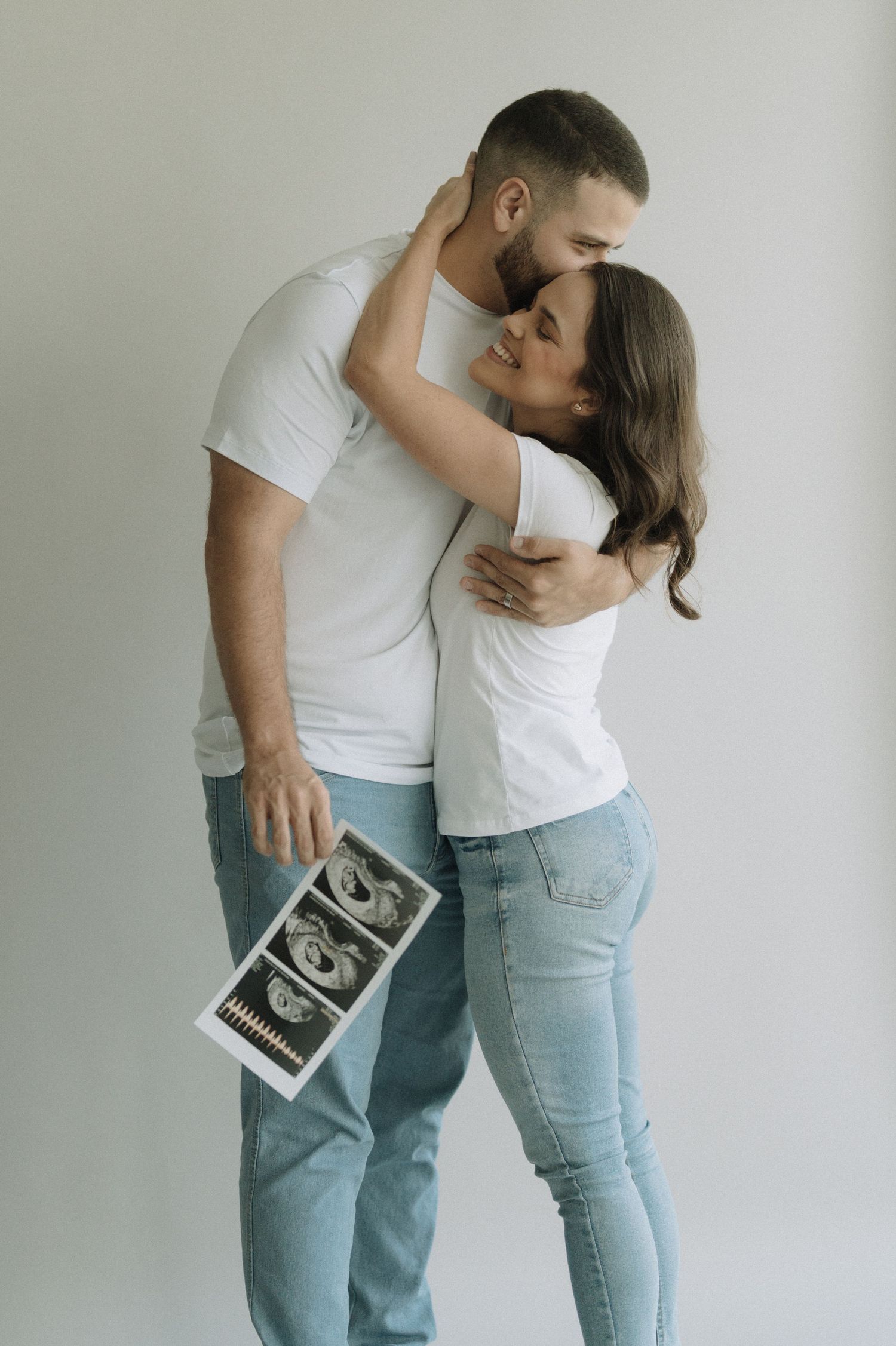 A couple in matching white tops and jeans embraces while holding an ultrasound photo in a minimalist setting.