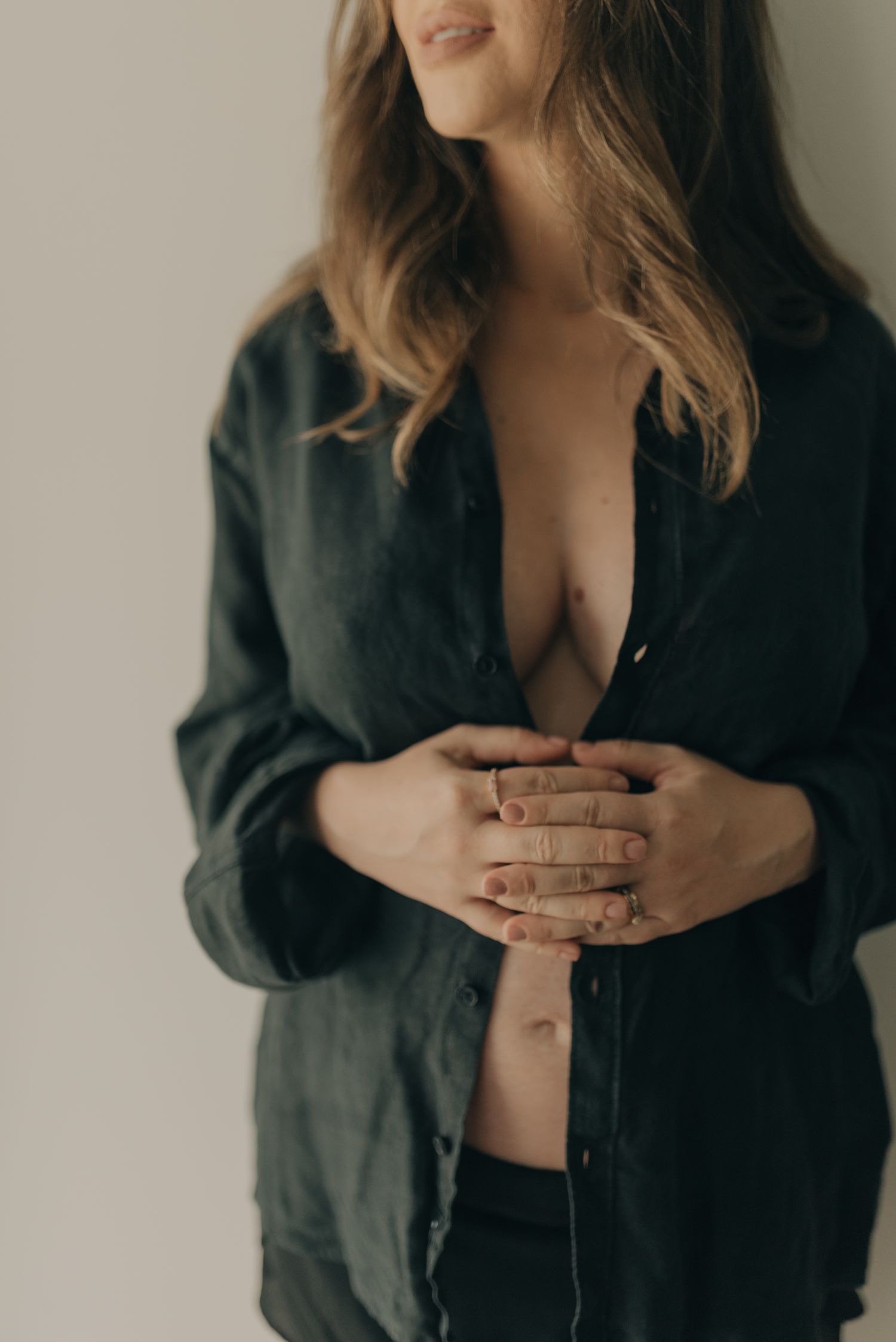 Intimate indoor portrait showing figure in oversized black button-down shirt against neutral wall.