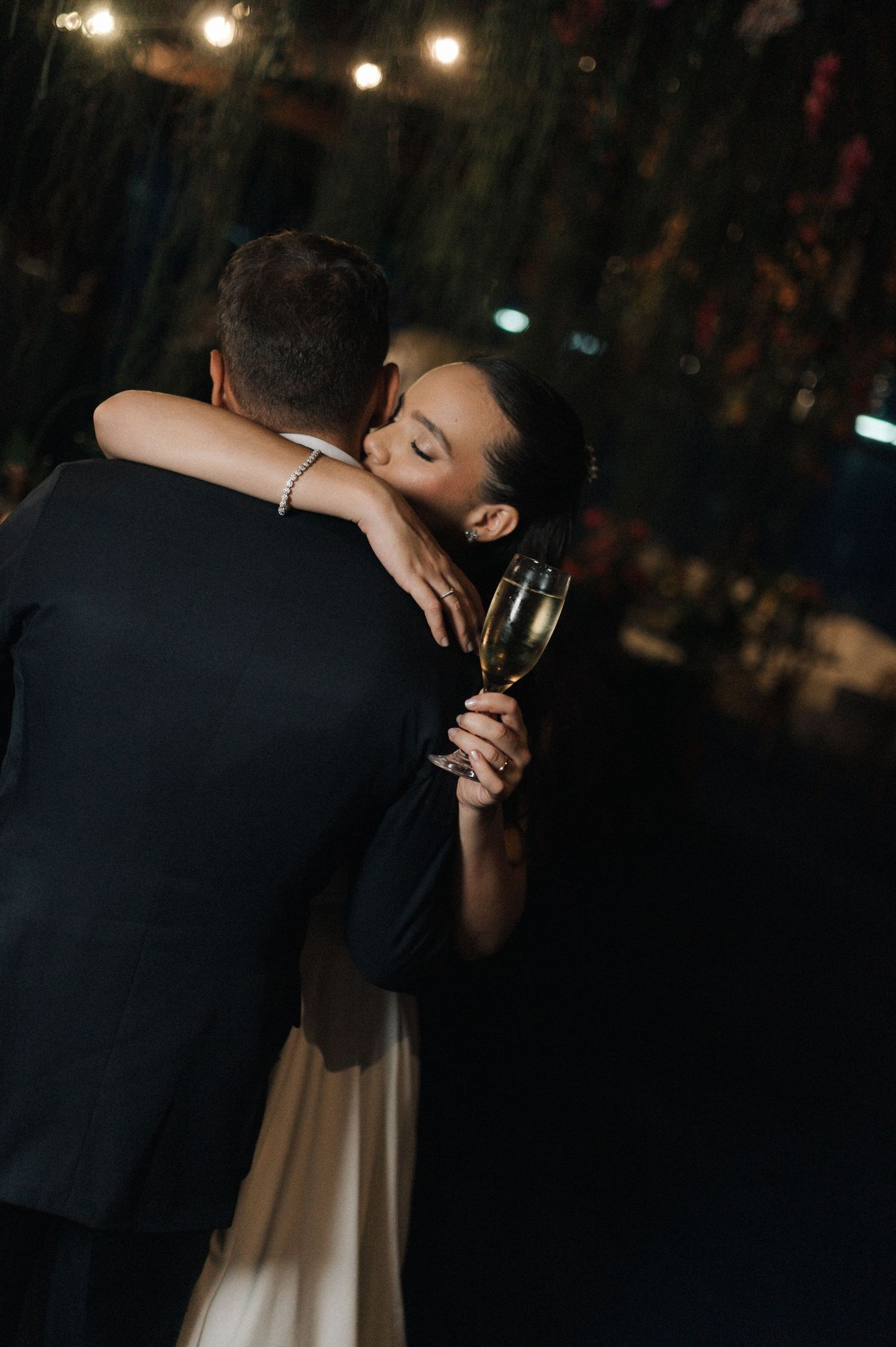 Bride holding champagne glass embraces groom during romantic first dance at elegant evening wedding reception.
