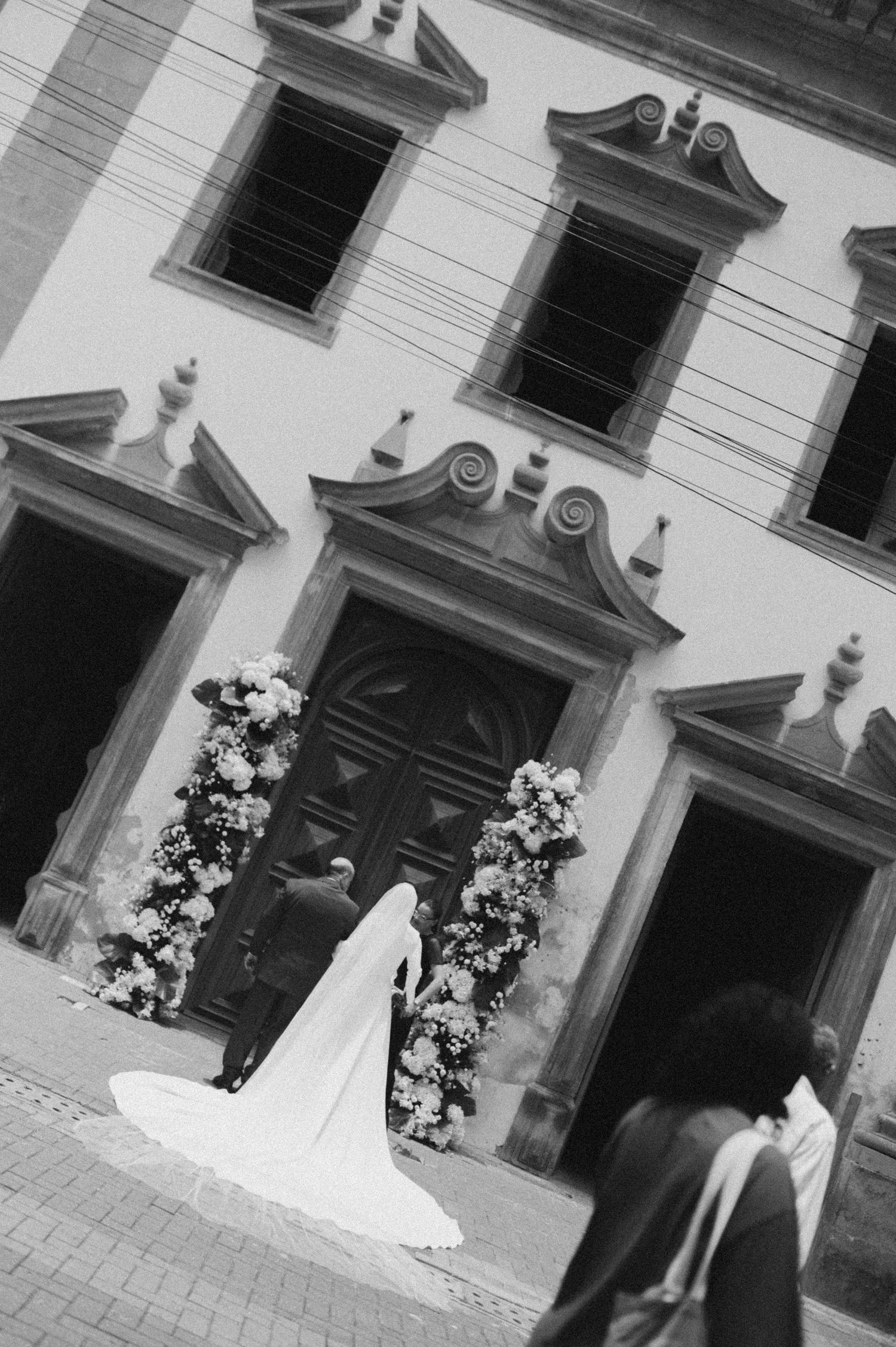 Black and white photo of bride in cathedral-length veil entering flower-adorned historic church doors.