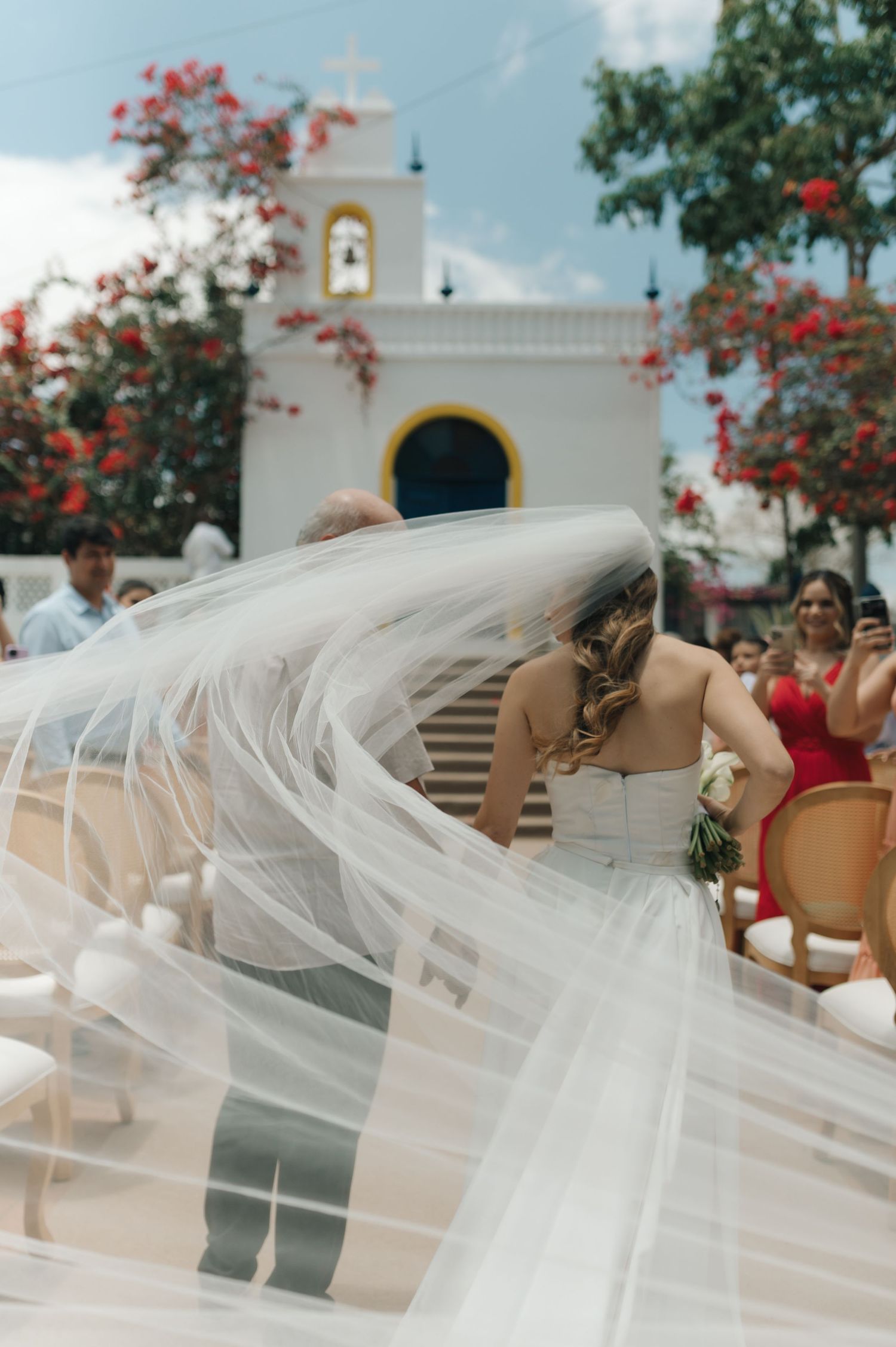 A long flowing veil catches the breeze in front of a white church with red flowers.