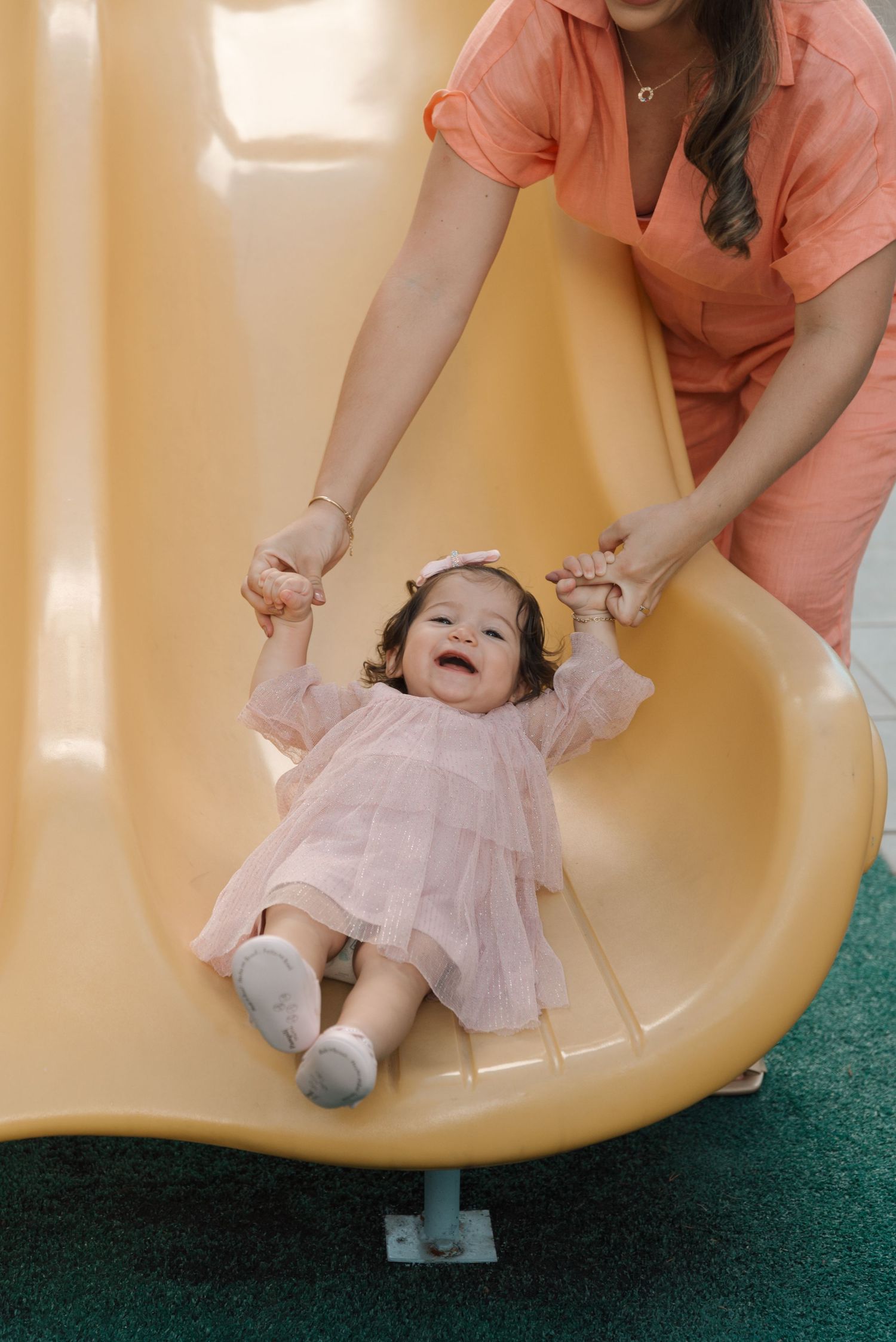 Toddler in pink dress laughs joyfully while being held on yellow playground slide.