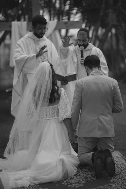 A wedding ceremony with priests blessing the kneeling couple outdoors.
