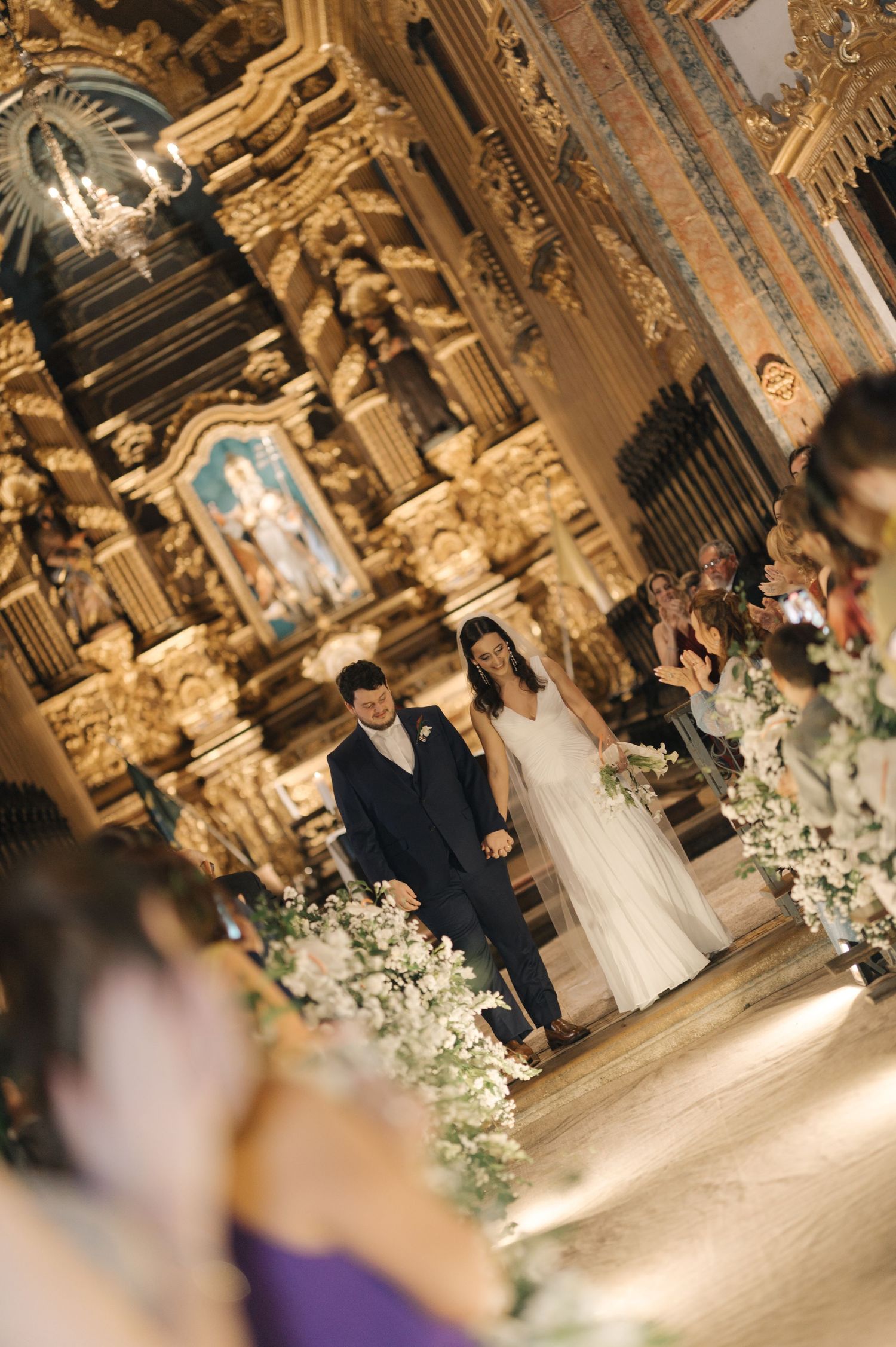 A traditional wedding ceremony takes place inside an ornate cathedral with baroque decor and white floral arrangements along the aisle.
