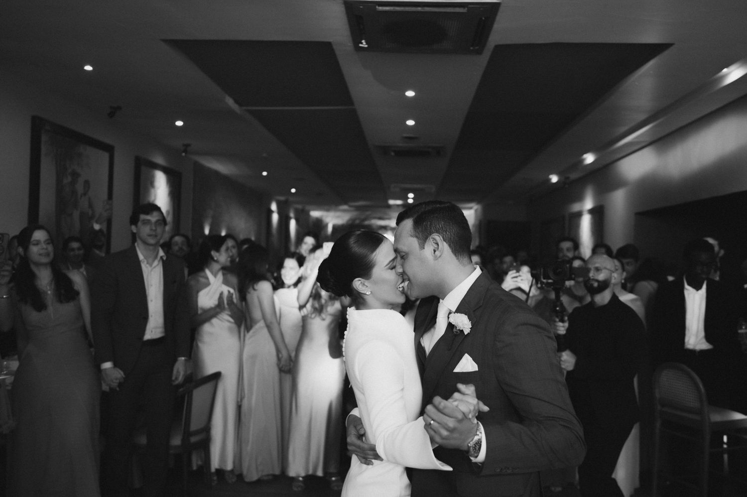 Black and white photo of a bride and groom sharing their first dance surrounded by guests at a wedding reception.