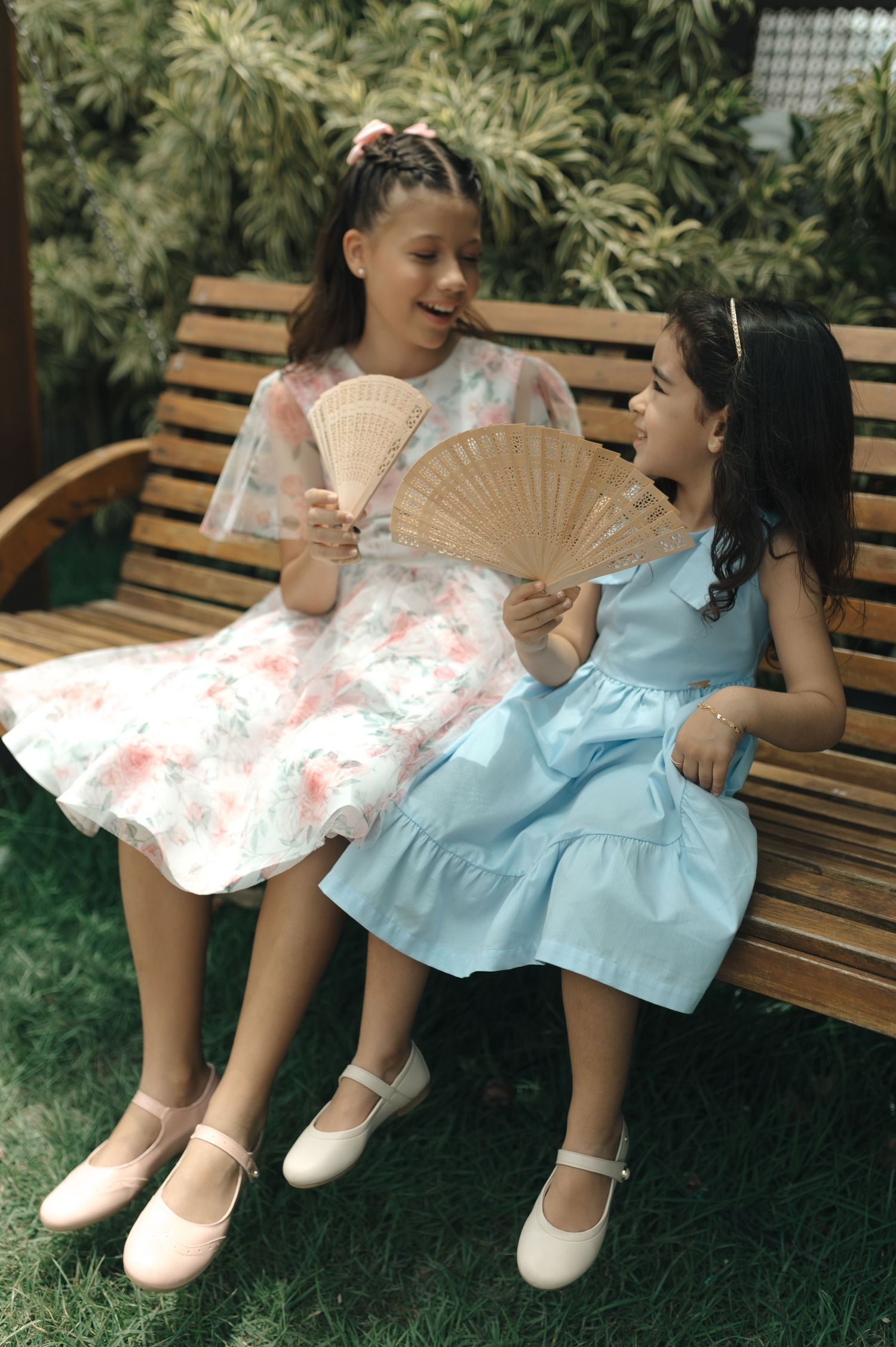 Two young girls in pretty dresses sitting on a garden bench, one holding a decorative fan.