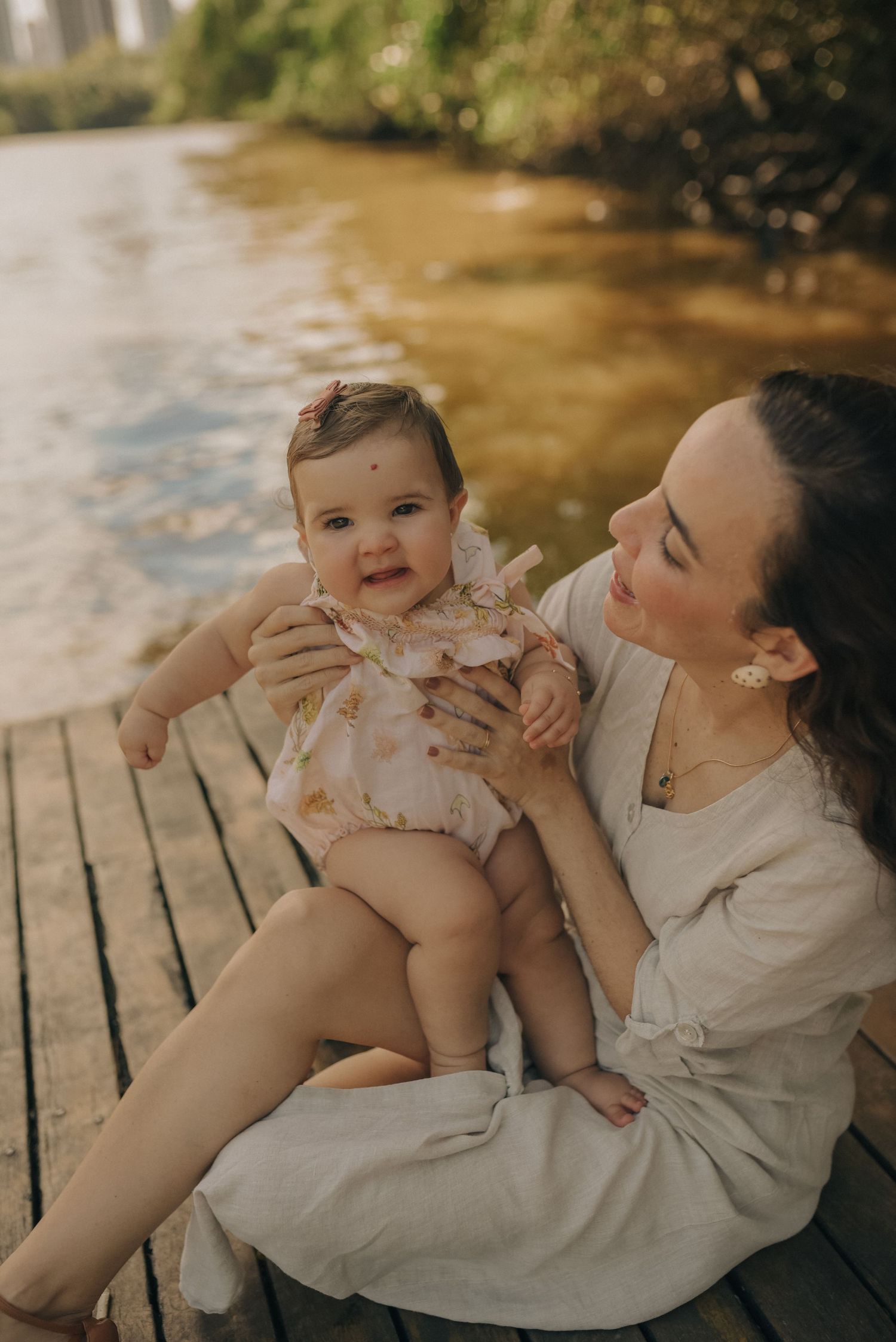 A tender moment between parent and baby sitting on a wooden dock by a sunlit river.