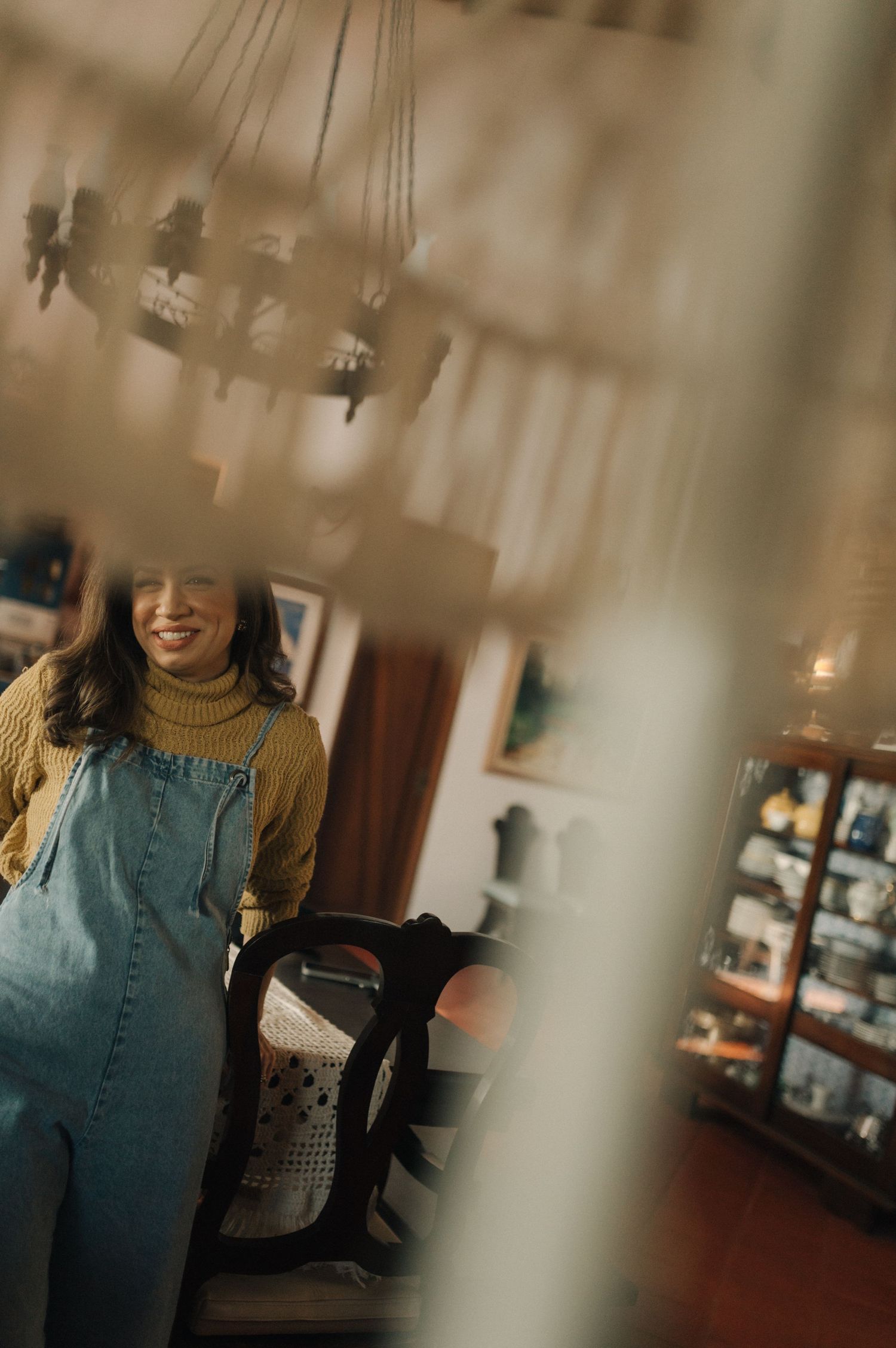 Artistic shot through hanging decorations showing someone in denim overalls and turtleneck.