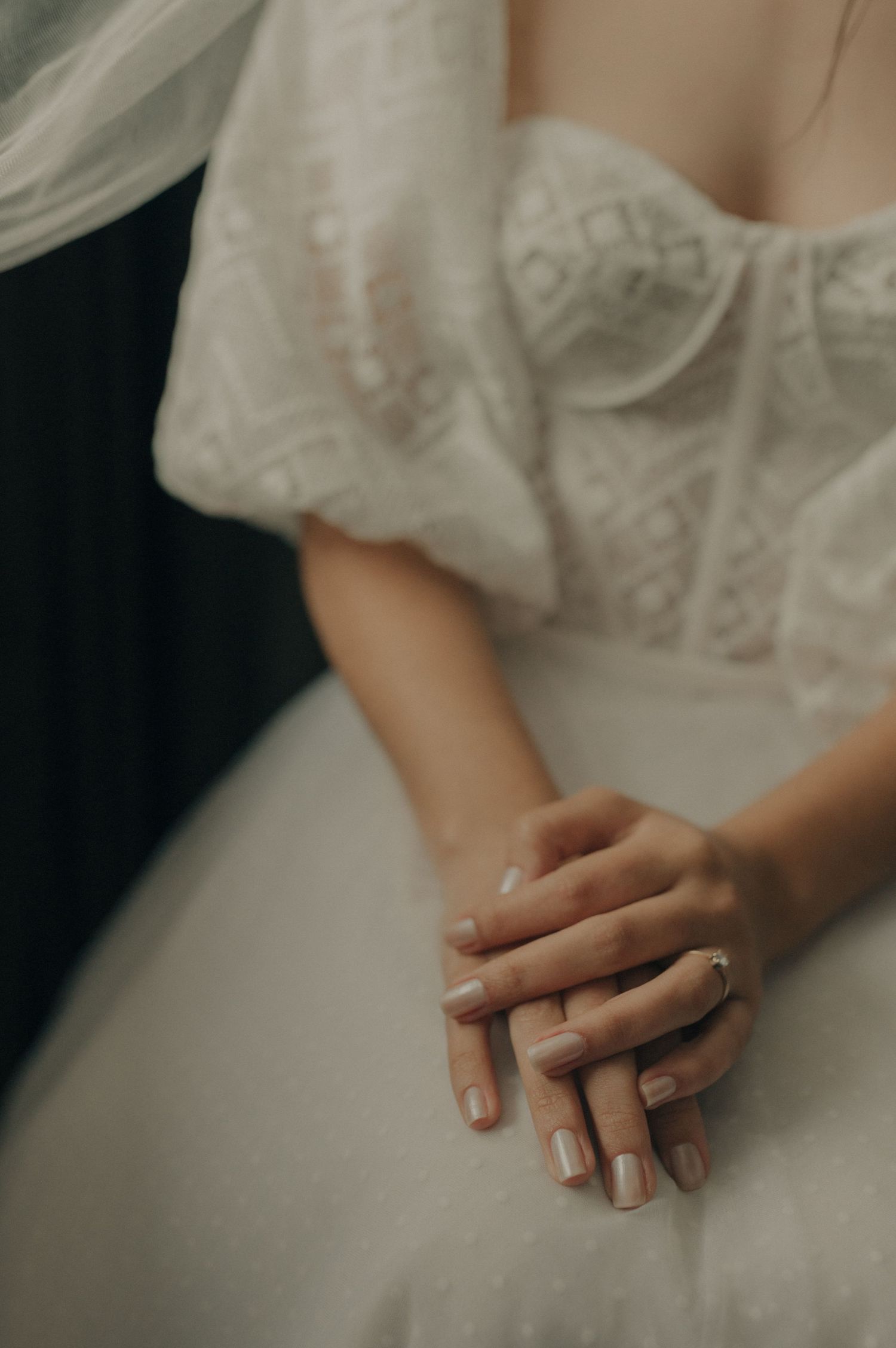 Close-up of folded hands resting on a white lace wedding dress.