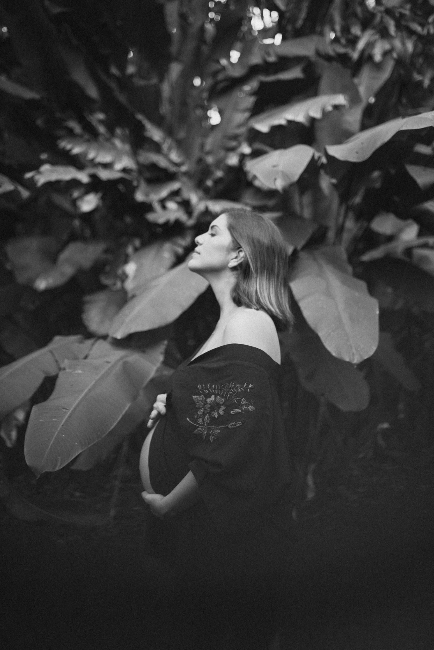 Artistic black and white maternity portrait among large tropical leaves in garden setting.