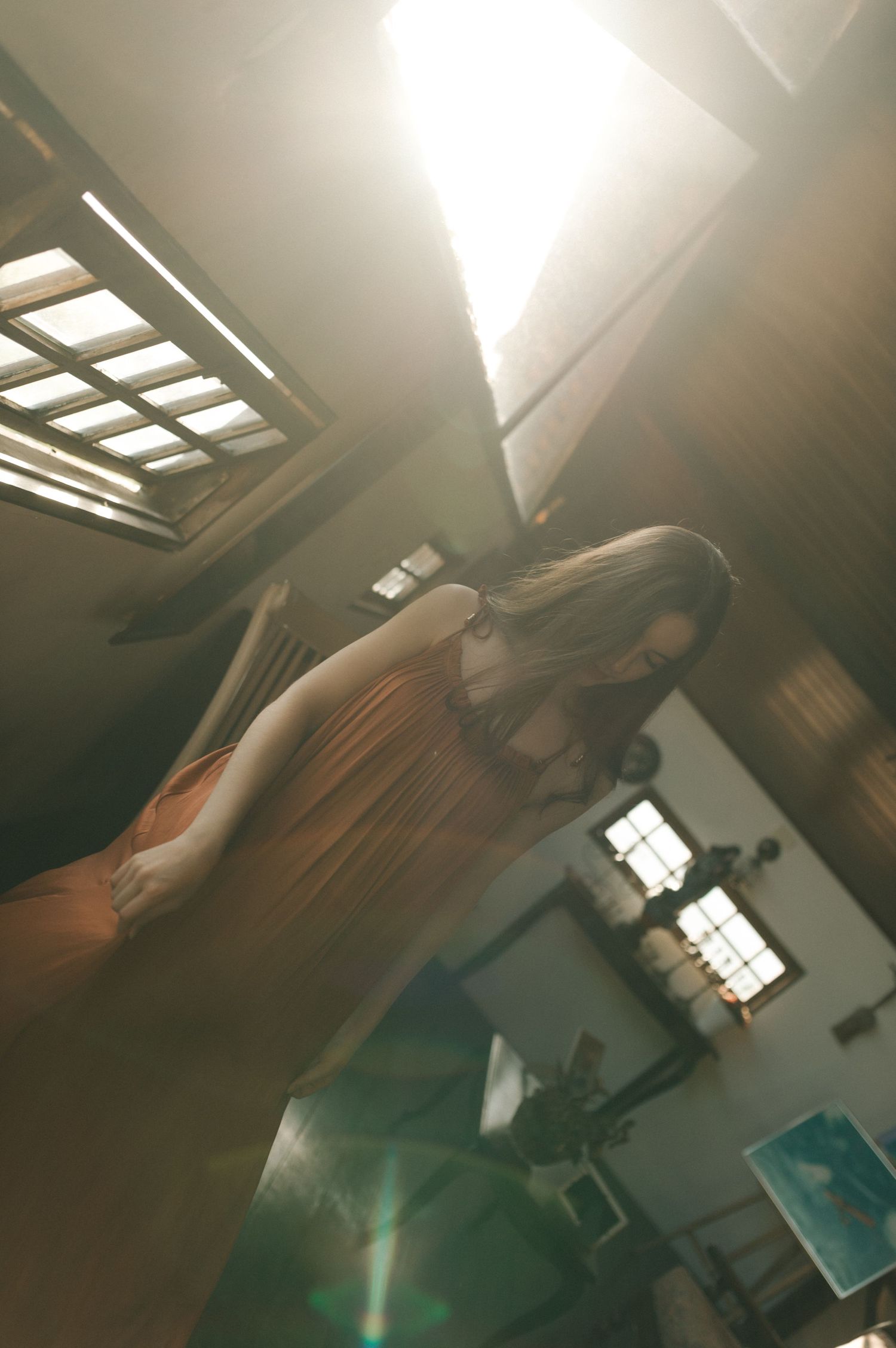 Interior shot of a figure in an orange dress standing beneath skylights with sunbeams streaming in.