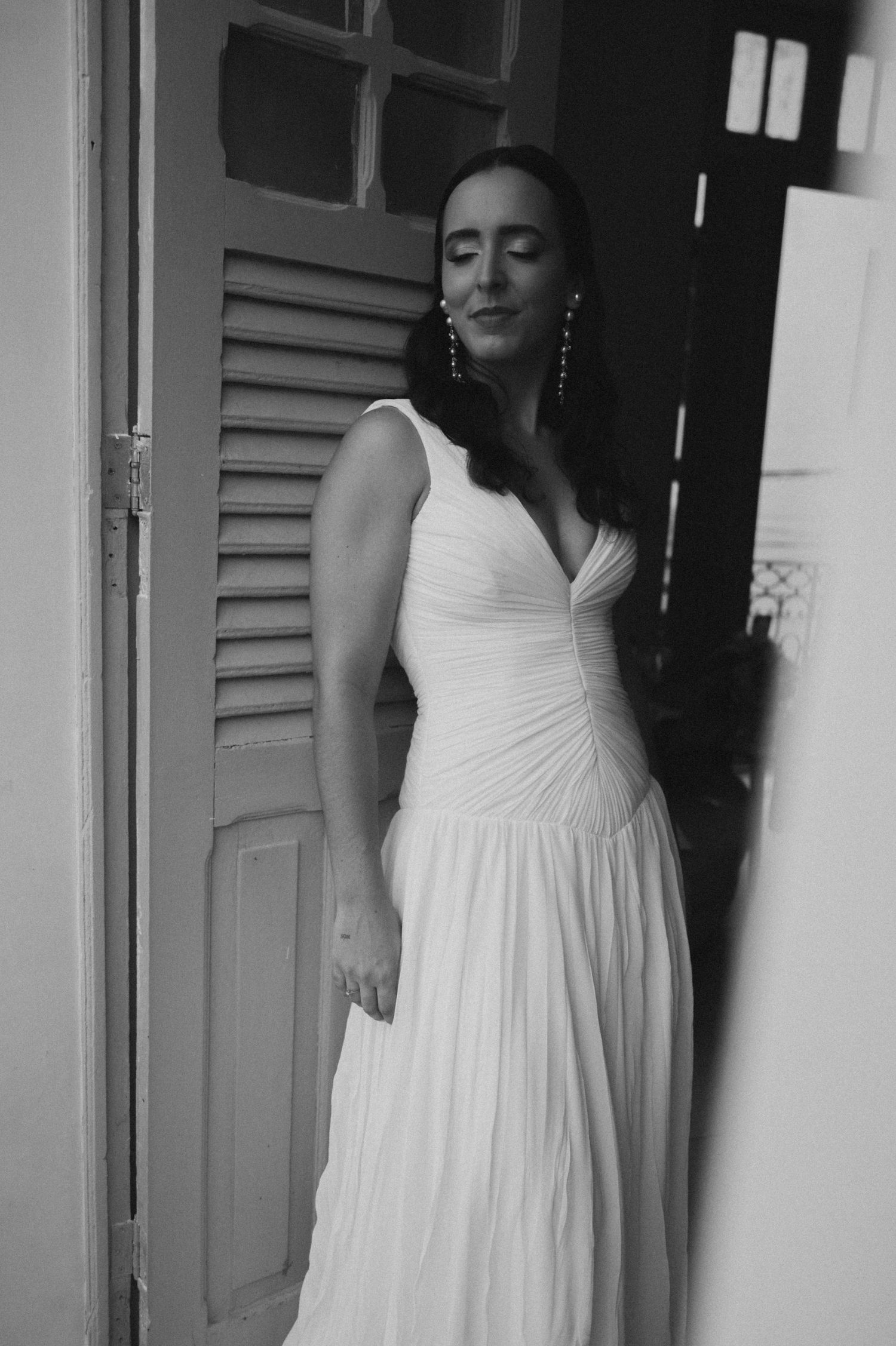 A person in a flowing white gown poses against a doorway in an elegant black and white photograph.