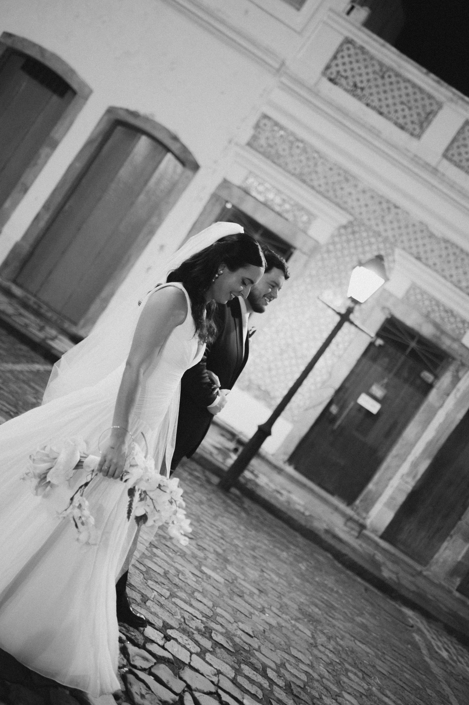 A romantic black and white wedding photo showing a couple in formal attire leaning together on a cobblestone street.