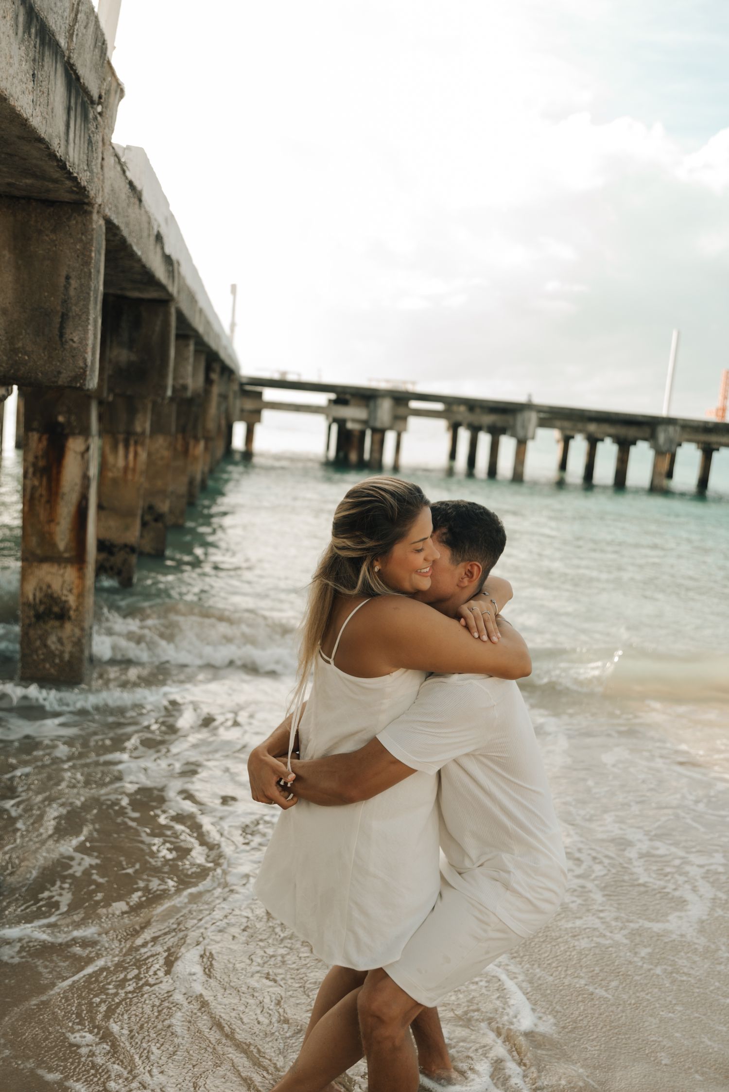 Couple in white clothing hugging and laughing in ocean waves near wooden pier at sunset.