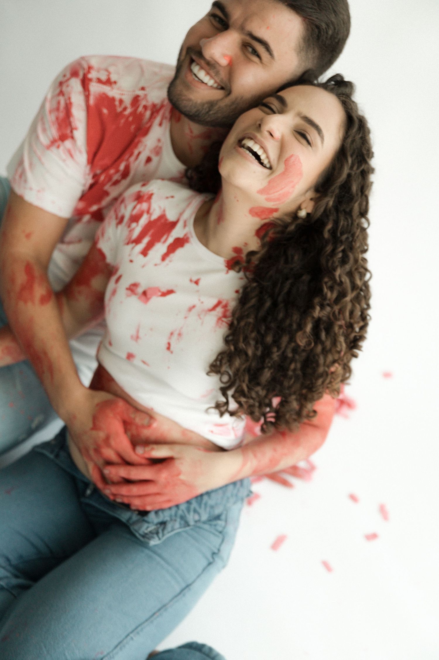 Happy couple laughing together, covered in red paint splatters, wearing white t-shirts and jeans.
