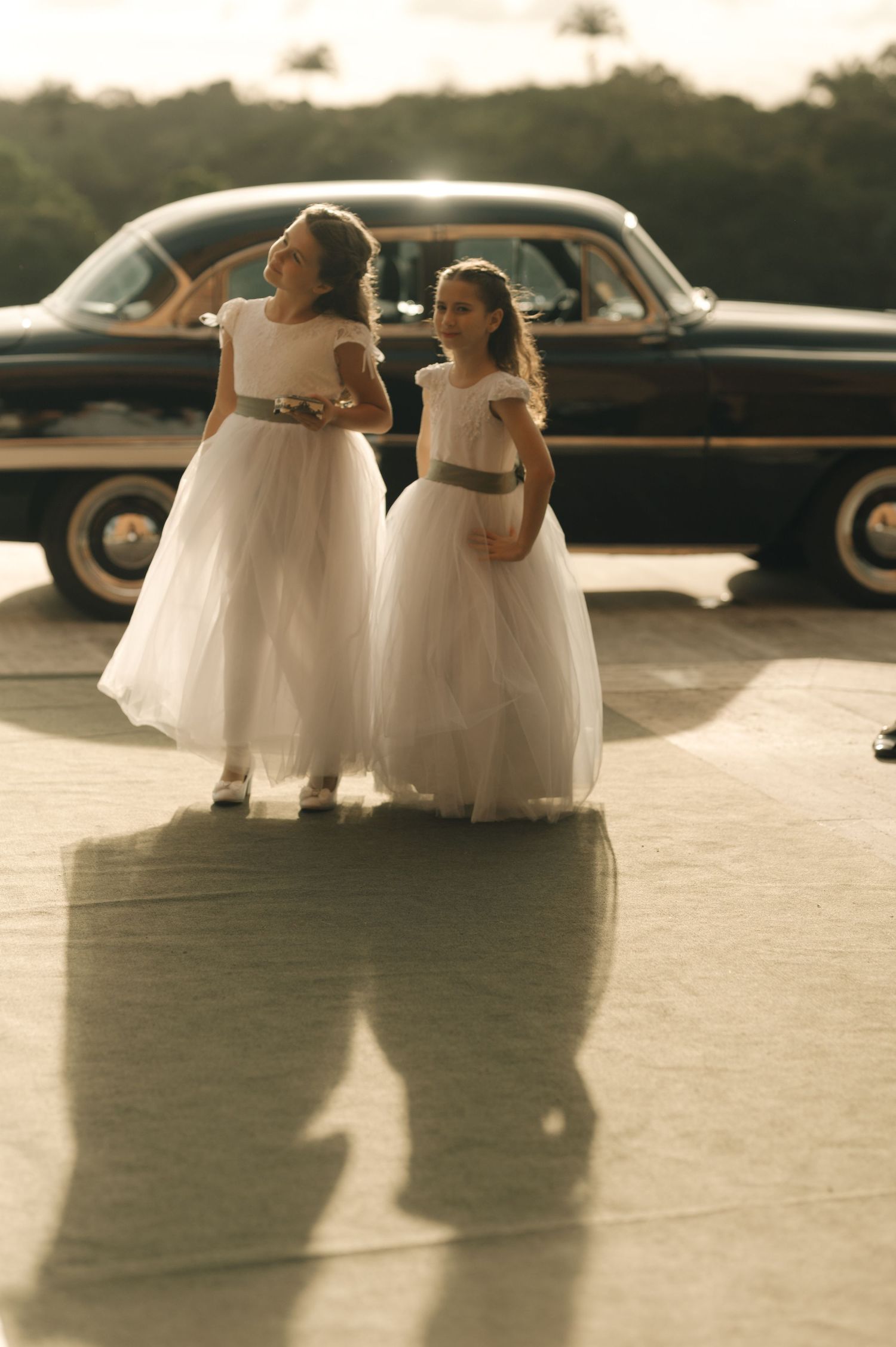 Two flower girls in white dresses walk beside a classic car at sunset casting long shadows on the ground.