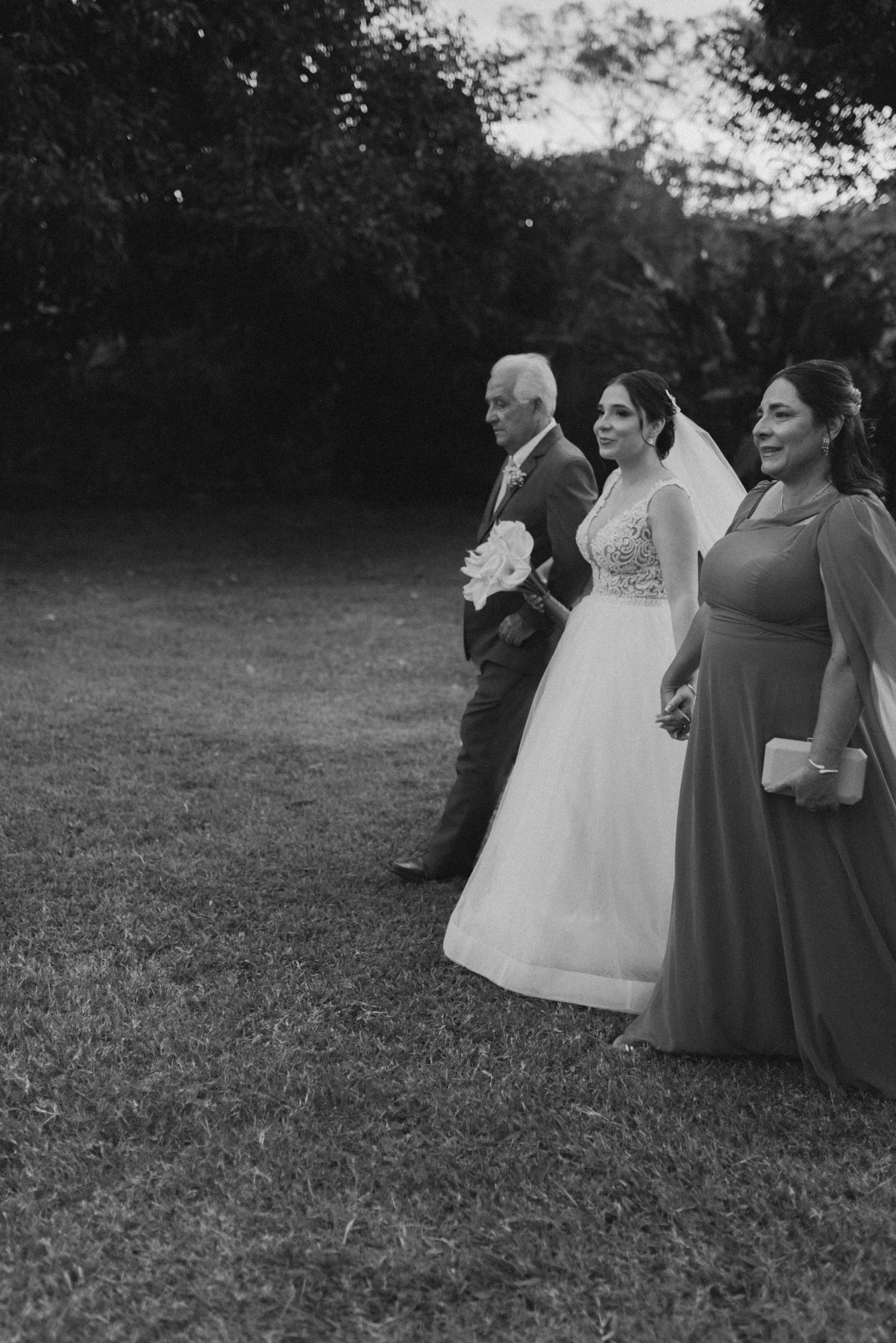 A wedding party in formal attire stands in a garden setting at dusk.