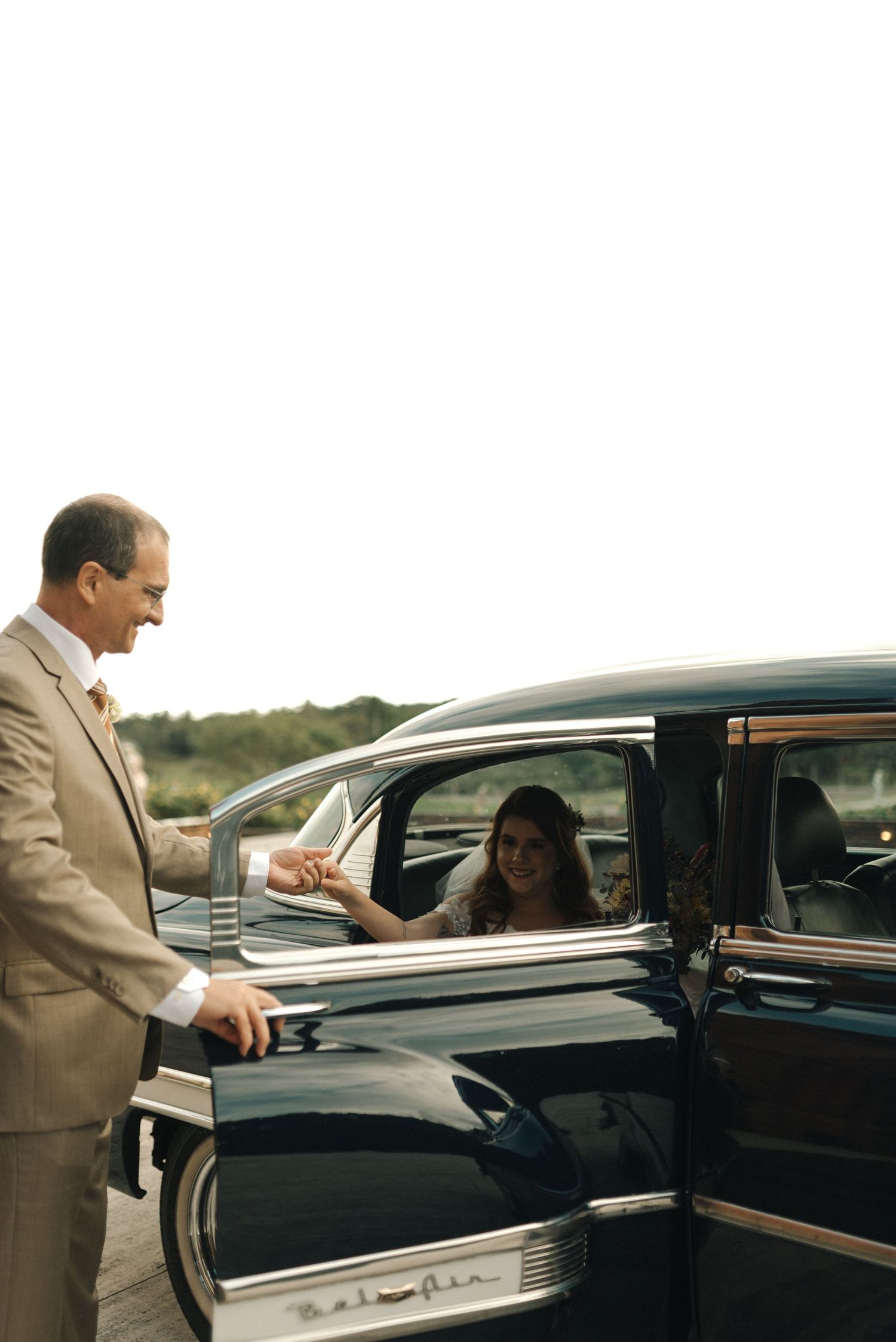A man in a beige suit stands beside a vintage black car, engaged in conversation through the driver's window.