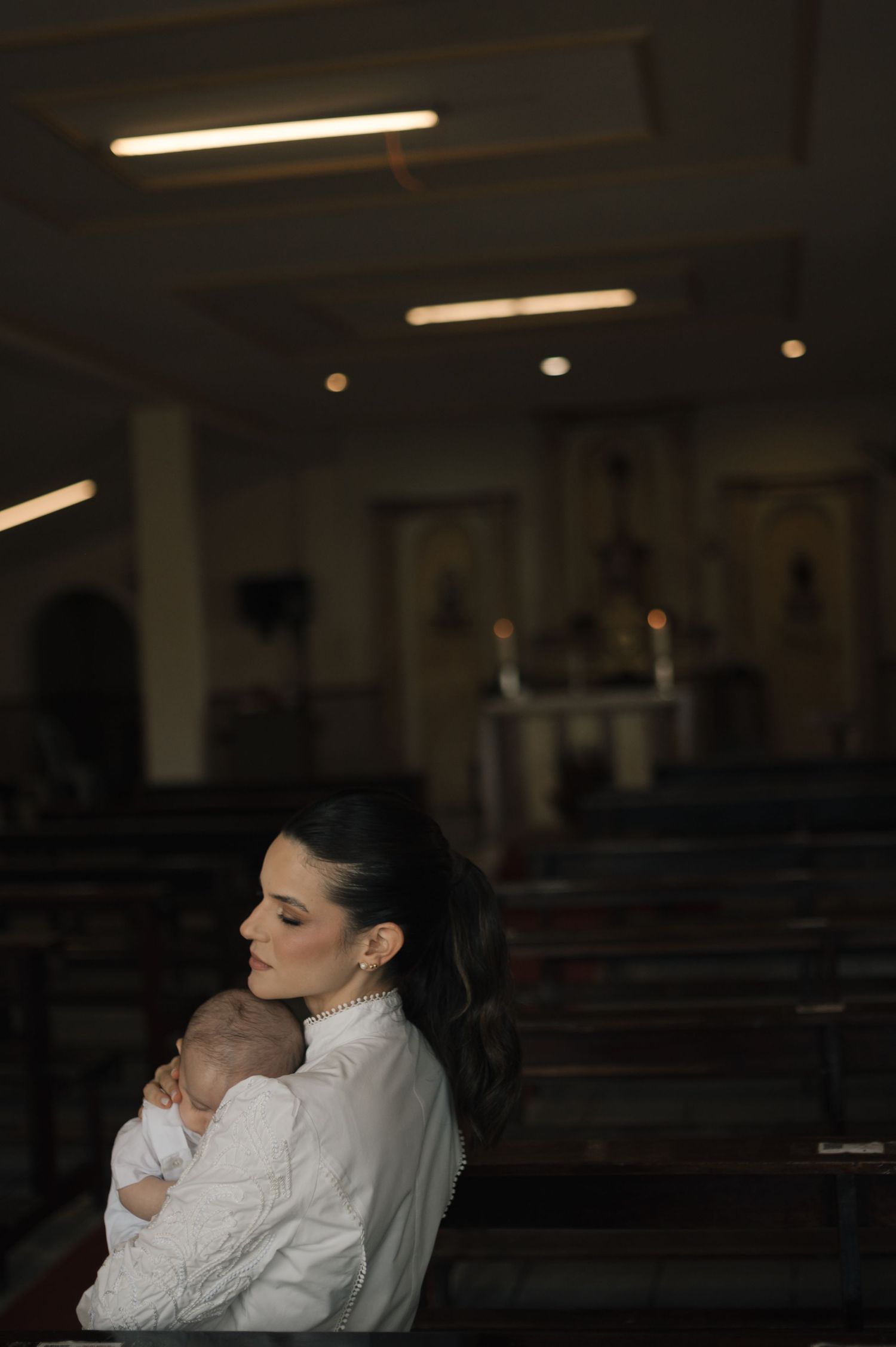 A woman in white holds a newborn baby in a dimly lit church, sitting peacefully in a pew.