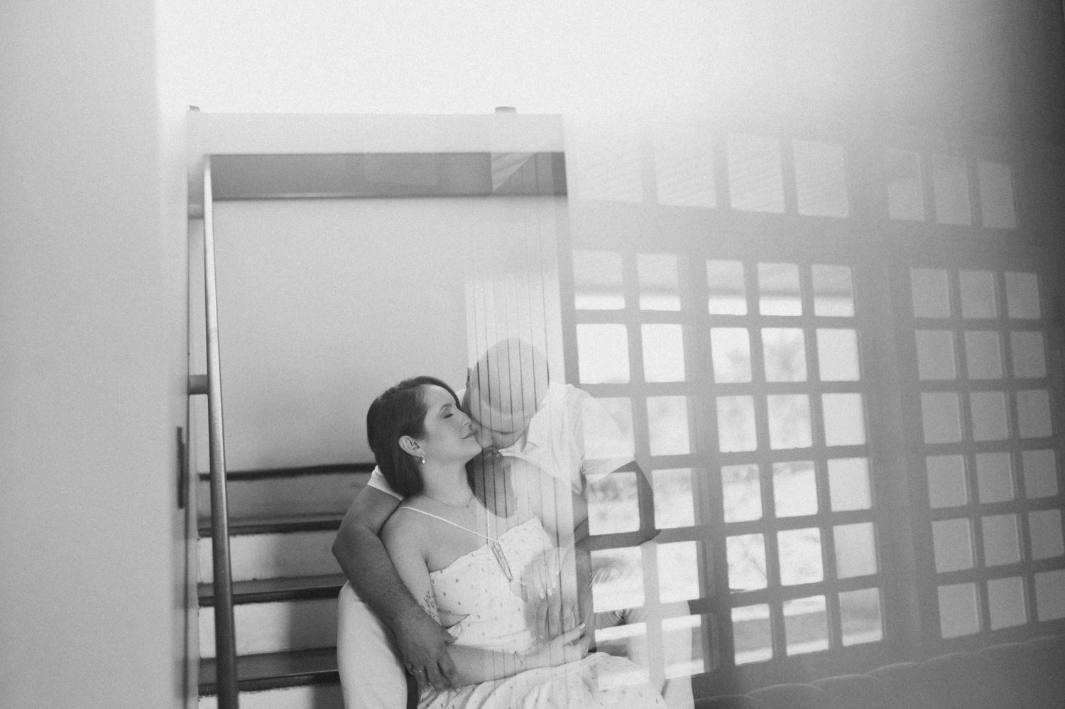 A couple in white dress clothes embracing against a gridded window.