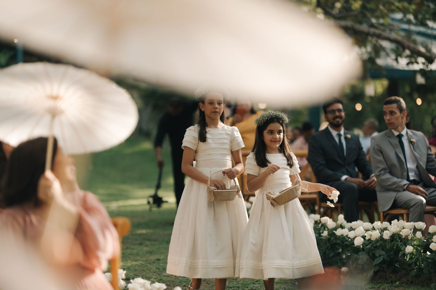 Two young attendants in white dresses walk down an outdoor aisle during a wedding ceremony.