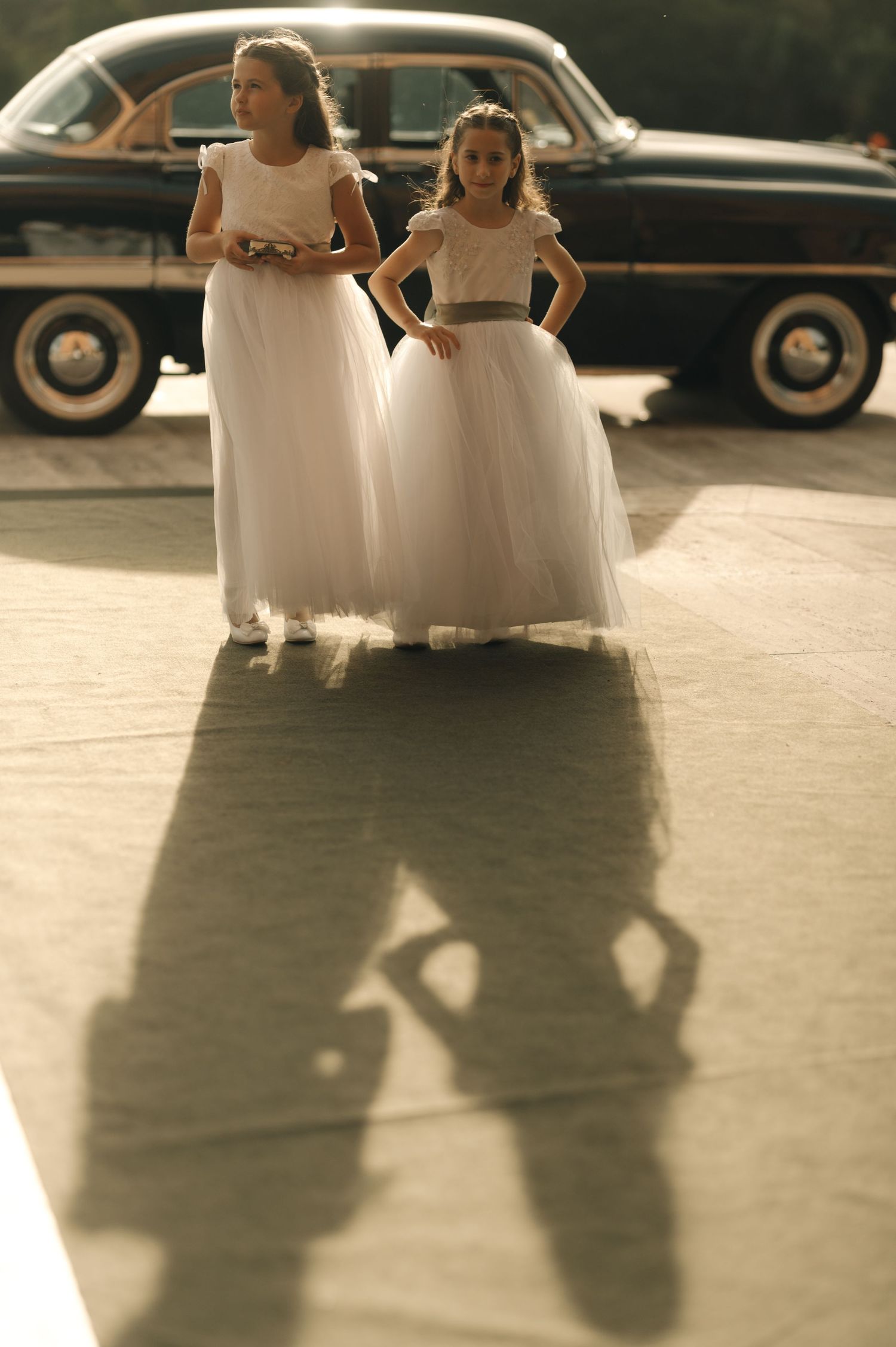 Two figures in white dresses cast long shadows on the ground while standing next to a vintage car.