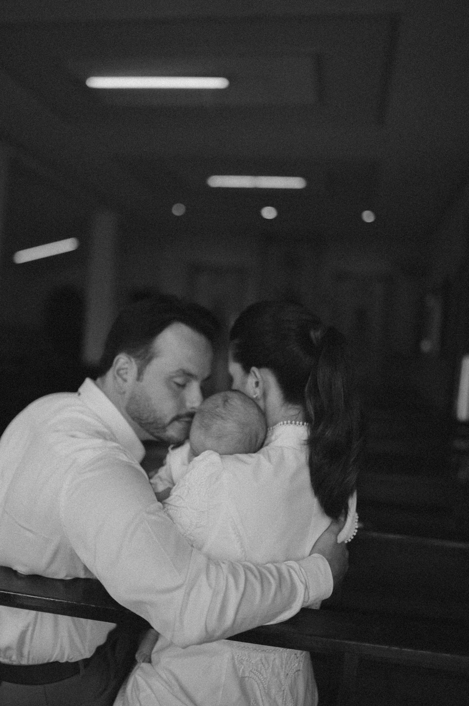 Black and white photo of parents sharing a tender moment kissing their newborn baby in a church setting.