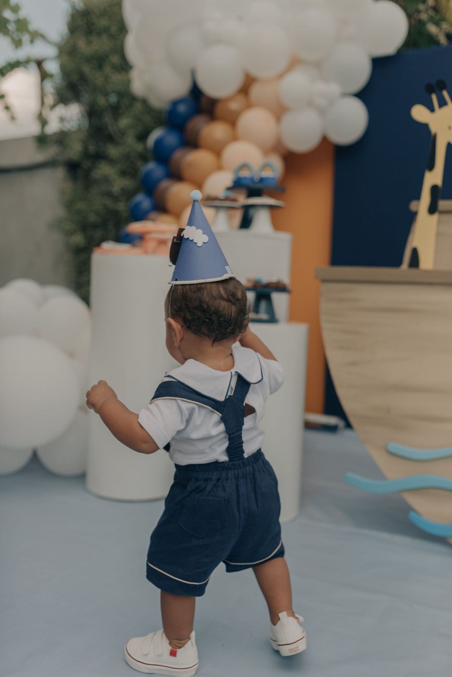 Small child in navy sailor outfit and party hat stands amid balloon decorations at celebration event.