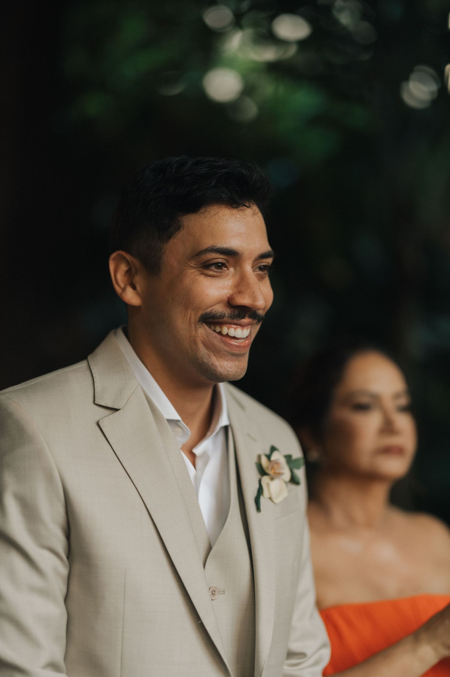 A wedding guest in a light beige suit smiles warmly during an outdoor celebration.
