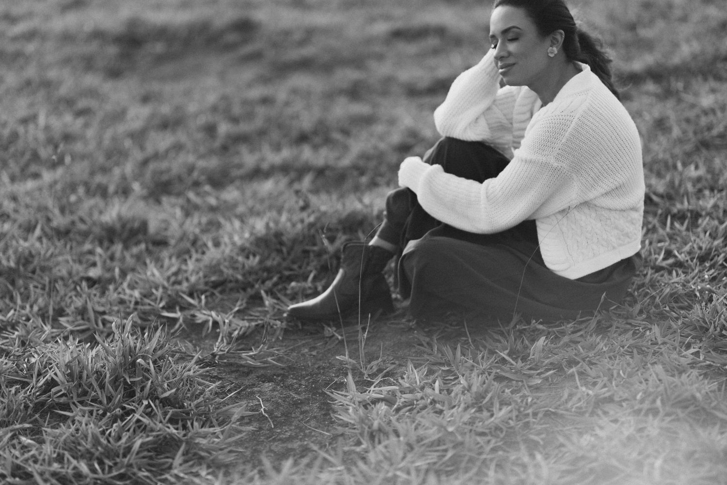 Black and white image of person sitting in grass field wearing white sweater.