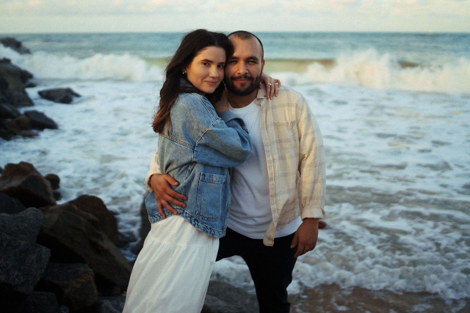 Romantic couple embracing on rocky beach at sunset with crashing waves in background.