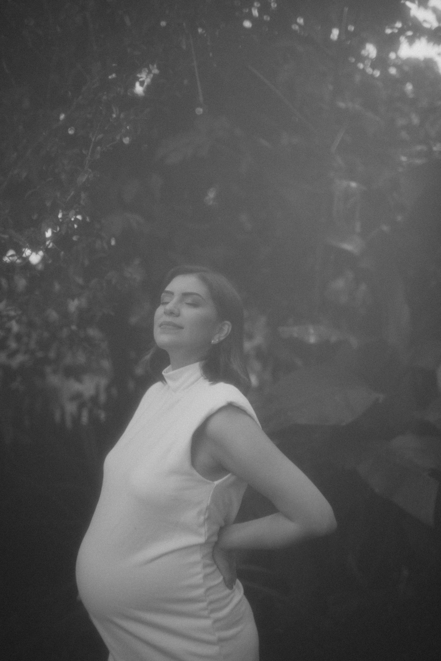Black and white maternity photo showing pregnant figure in white sleeveless top against natural backdrop.