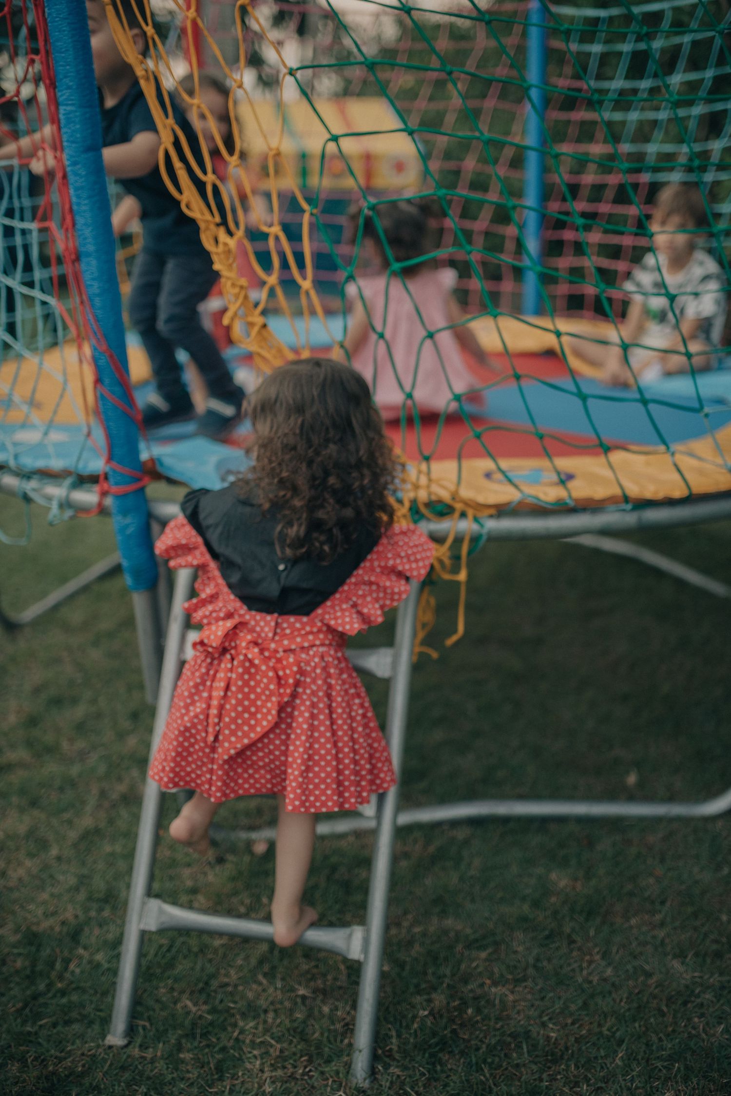 Child climbing ladder near blue netting outdoors in polka dot dress.
