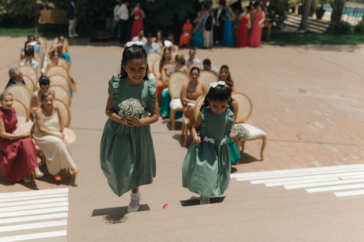 Two young attendants in teal dresses walk down an aisle during a wedding ceremony.
