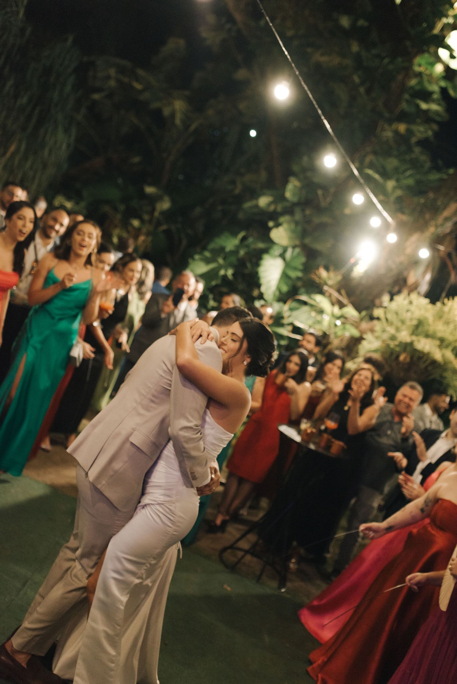 A couple shares their first dance at a nighttime wedding reception under string lights while guests watch.