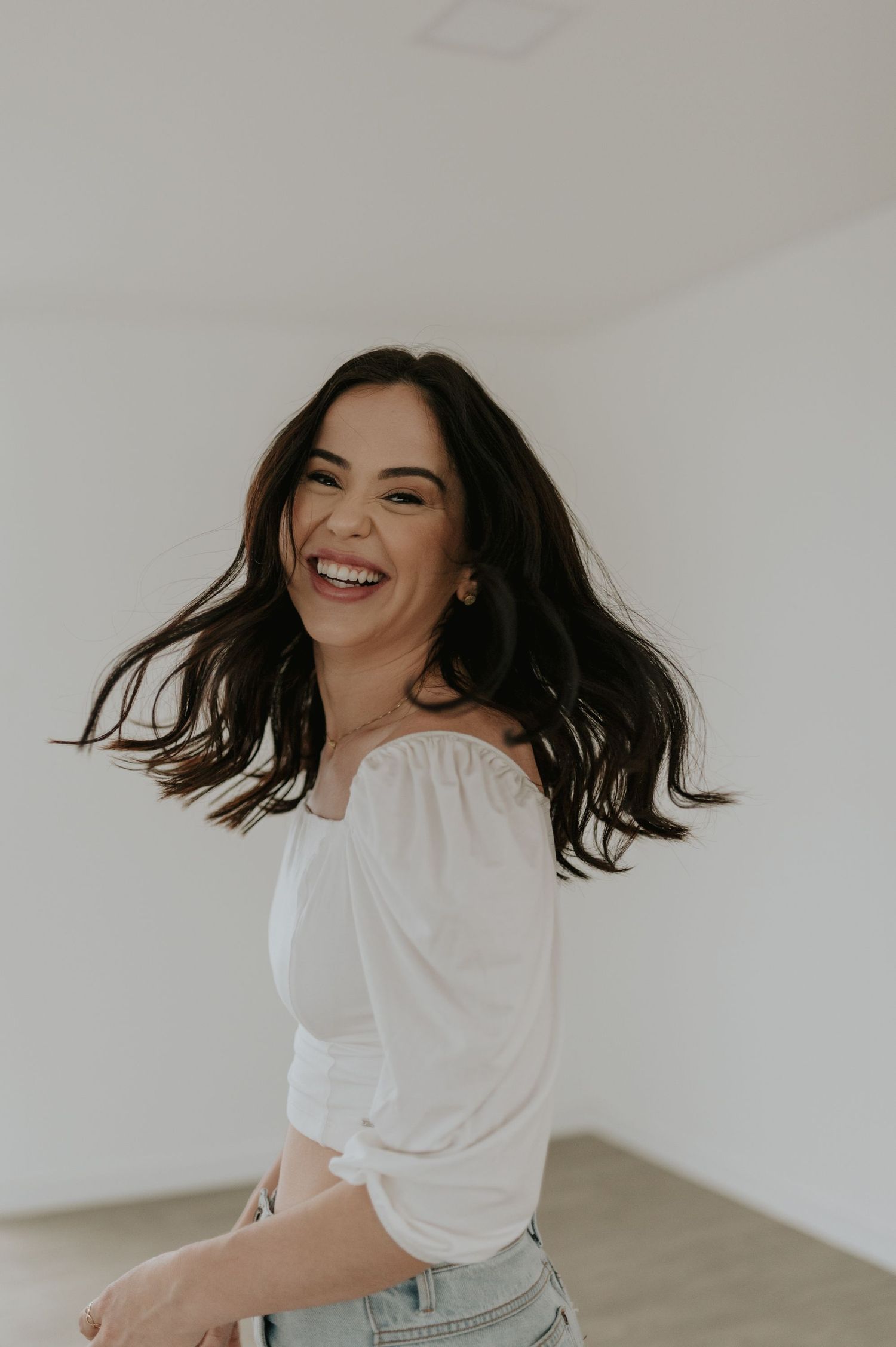 Candid portrait of woman with dark hair laughing in white blouse against minimal background.