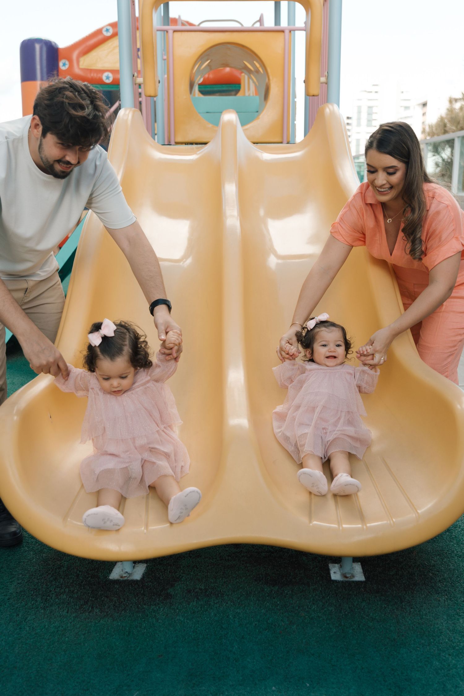 Parents helping toddlers slide down yellow playground slide together.