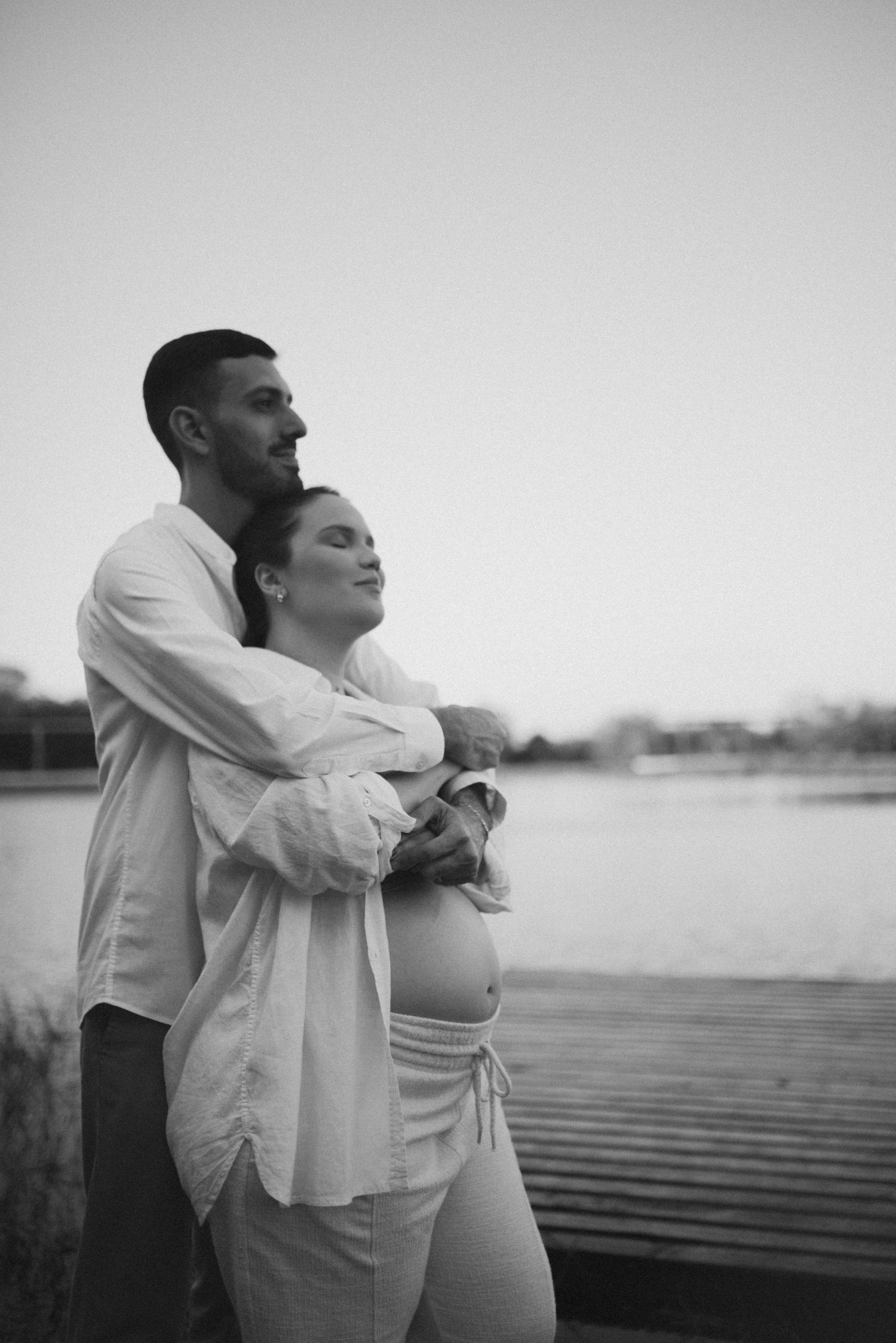 An intimate black and white maternity photo taken on a wooden dock overlooking calm water.