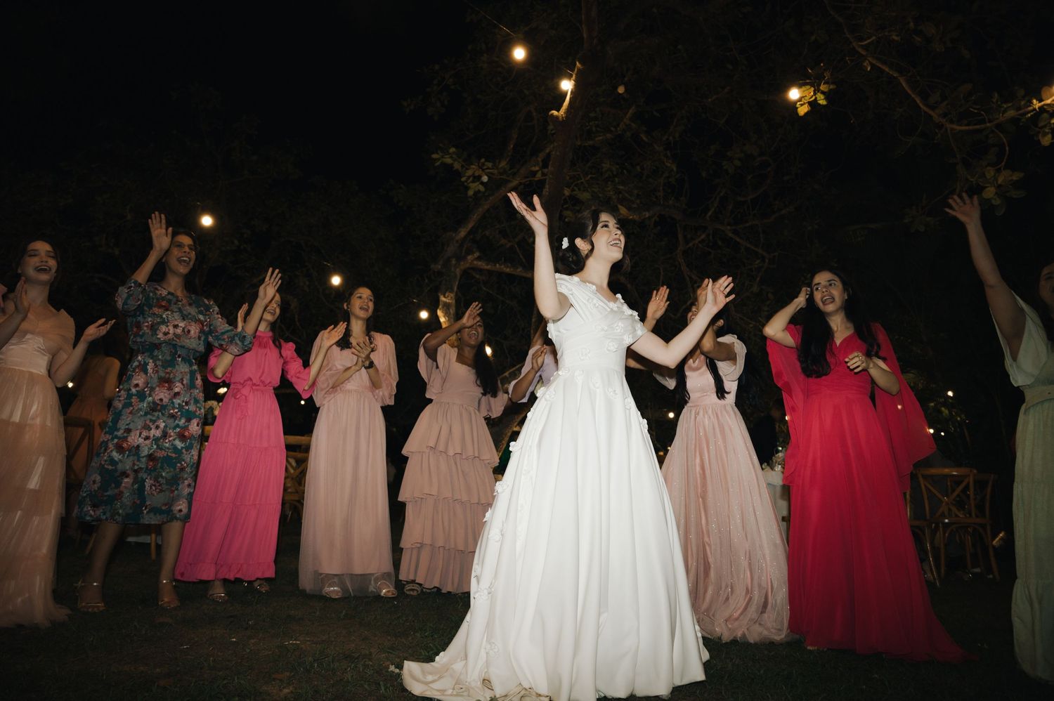 A joyous celebration with wedding party members dancing outdoors at night under string lights.
