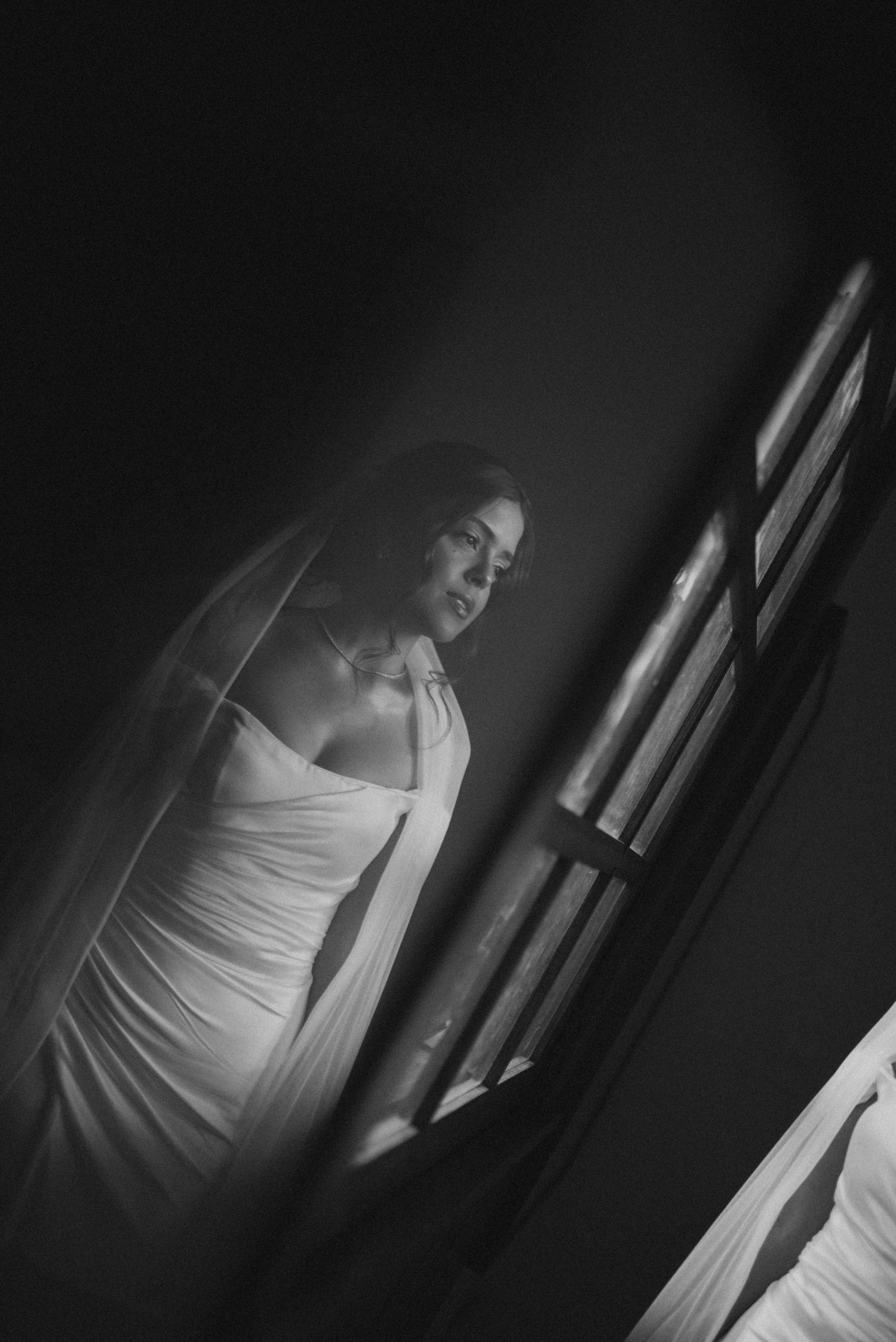A bride in an elegant white dress poses by a window in dramatic black and white lighting.
