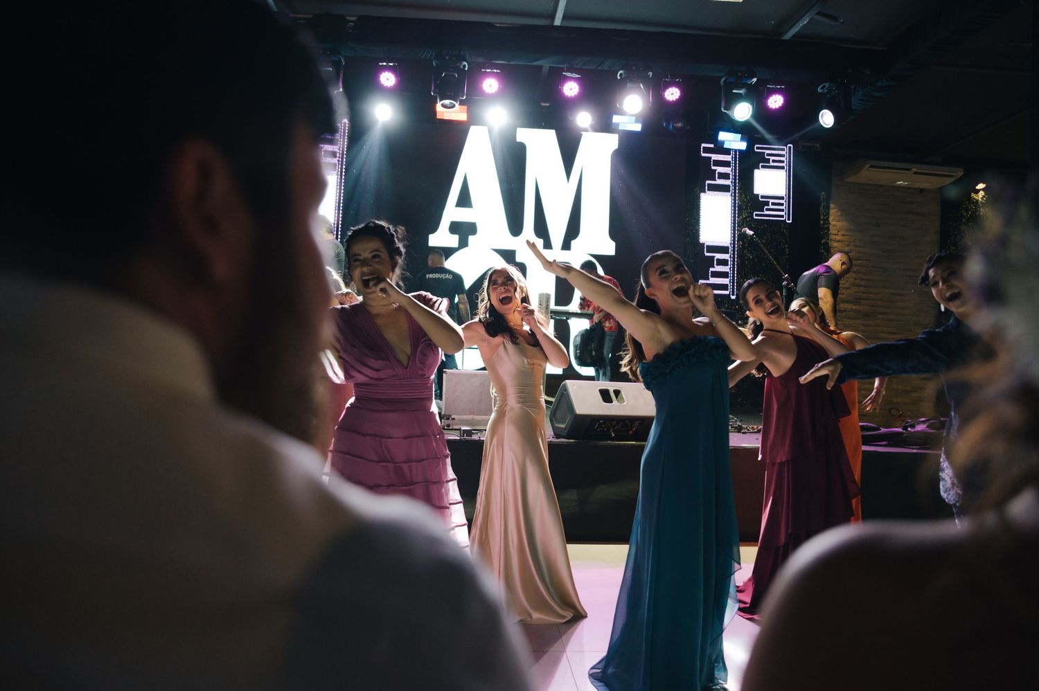 Wedding guests dance and celebrate on a dark dance floor with illuminated AM letters and stage lighting in the background.