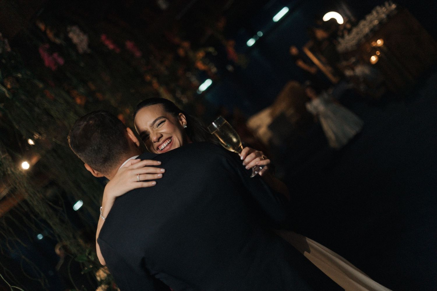 A smiling woman in elegant attire holds a champagne glass while embracing a man in a black suit at a formal event.