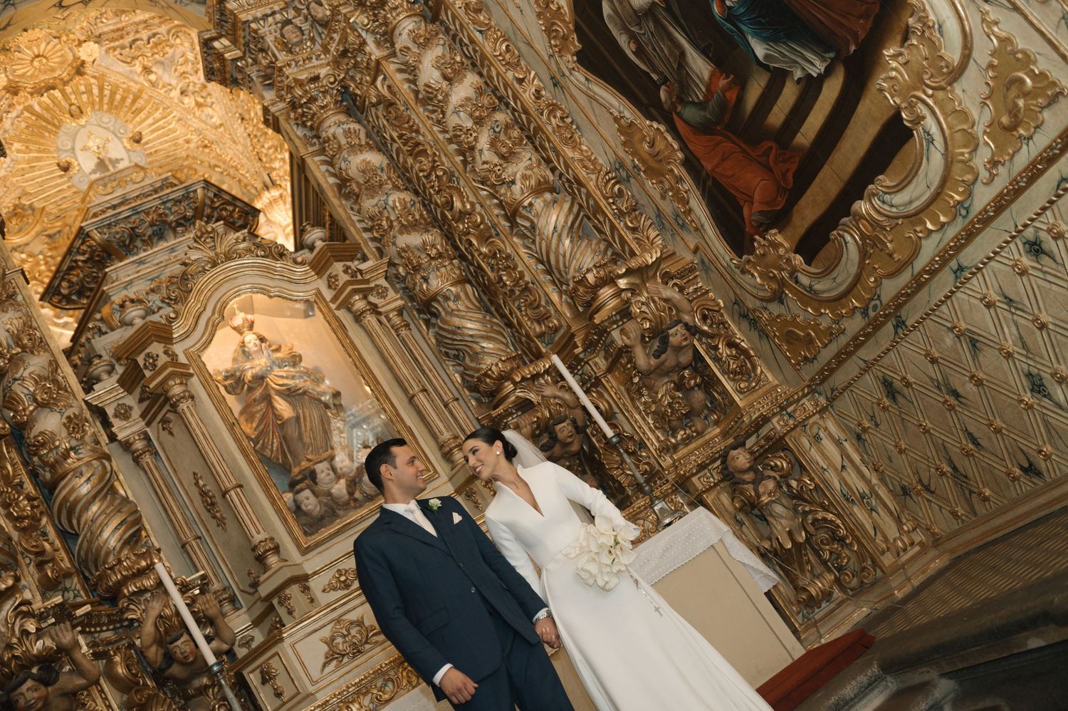 Bride and groom stand before an elaborate golden baroque altar inside a historic ornate church.