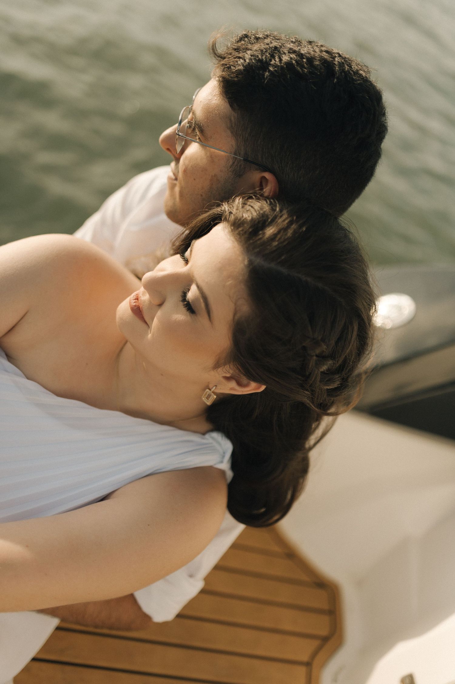 Romantic couple in white clothing relaxing together on a wooden deck by the water in warm sunlight.